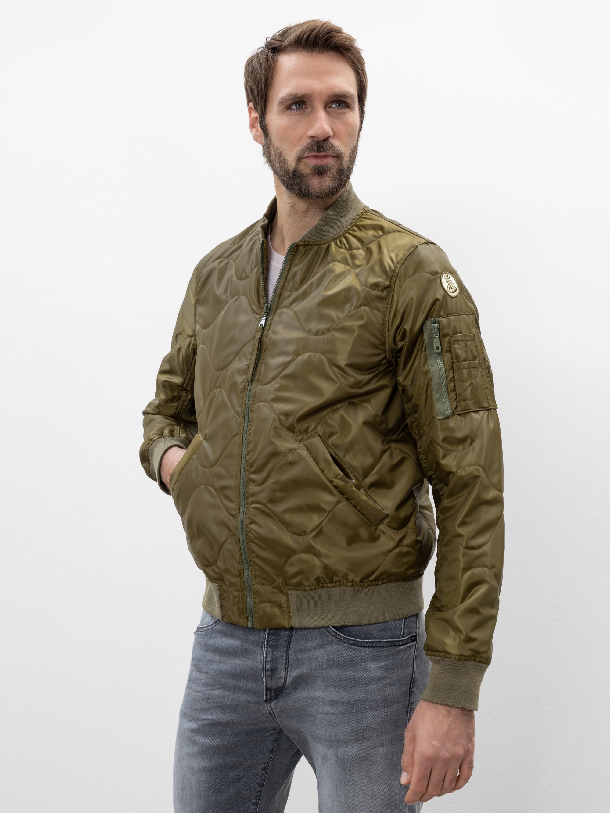 Ein Mann mit kurzen braunen Haaren und einem Bart trägt eine glänzende olivgrüne Bomberjacke und hellgraue Jeans. Er steht vor einem schlichten weißen Hintergrund und hat die Hände in den Jackentaschen.