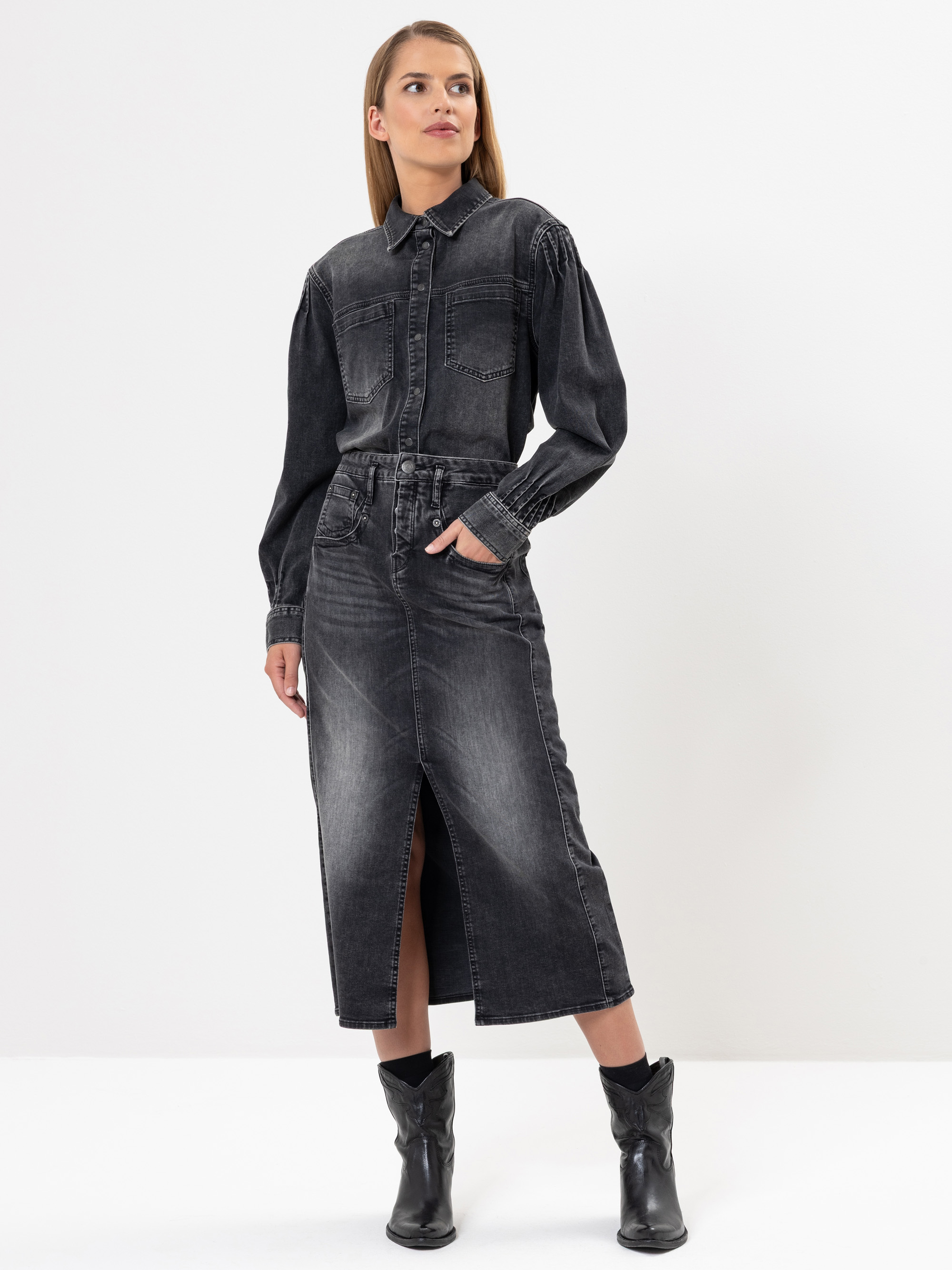 Eine Frau, die ein schwarzes Jeanshemd und einen dazu passenden schwarzen Jeans-Midirock mit einem vorderen Schlitz trägt, steht mit einer Hand in der Tasche und der anderen an ihrer Seite. Außerdem trägt sie schwarze Ankle Boots vor einem schlichten w