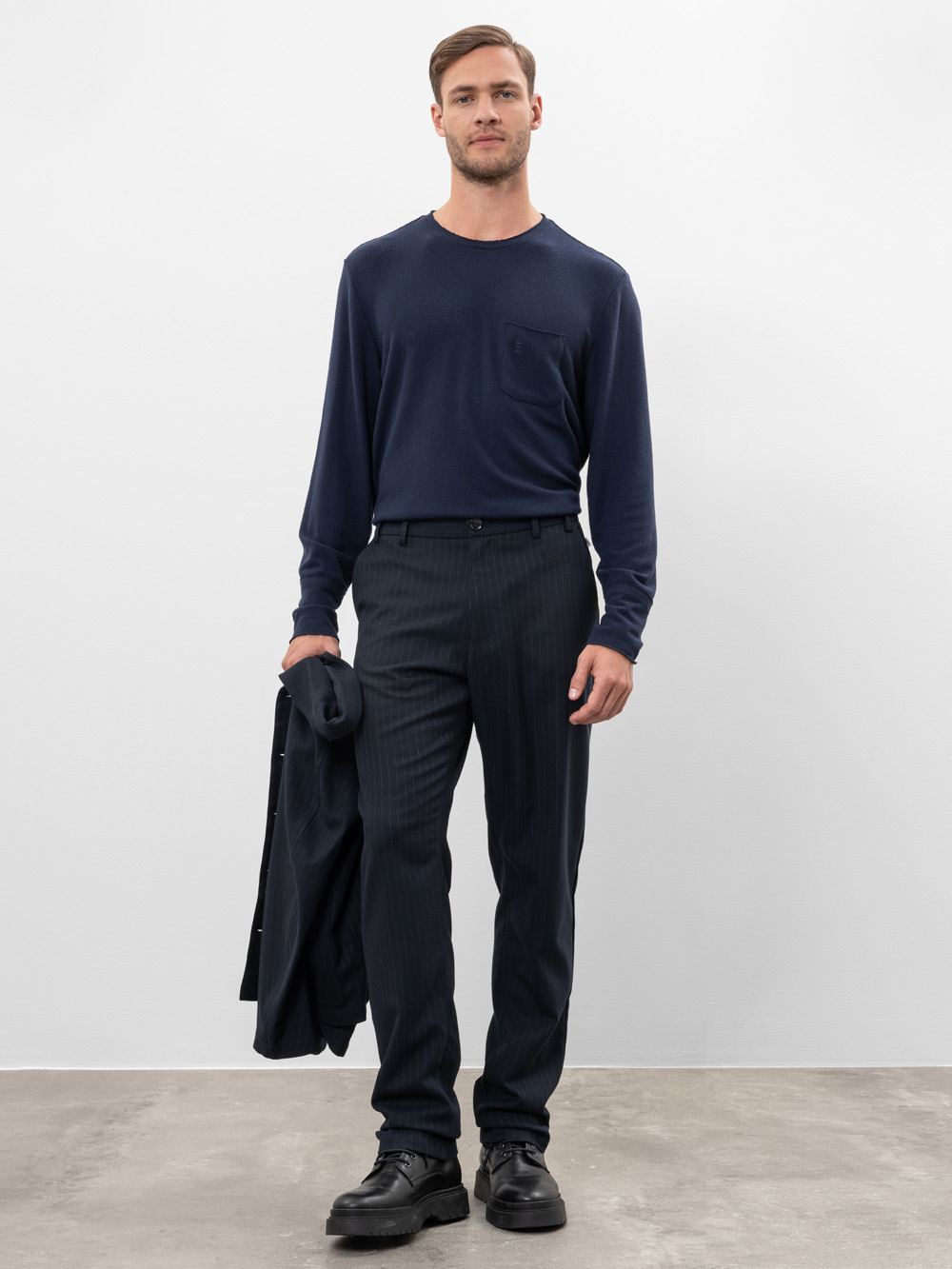 Ein Mann steht vor einem schlichten Hintergrund und trägt ein marineblaues langärmeliges Hemd, das in eine dunkle Nadelstreifenhose gesteckt ist. Er hält eine dunkle Jacke in einer Hand, trägt schwarze Schuhe und blickt direkt in die Kamera.