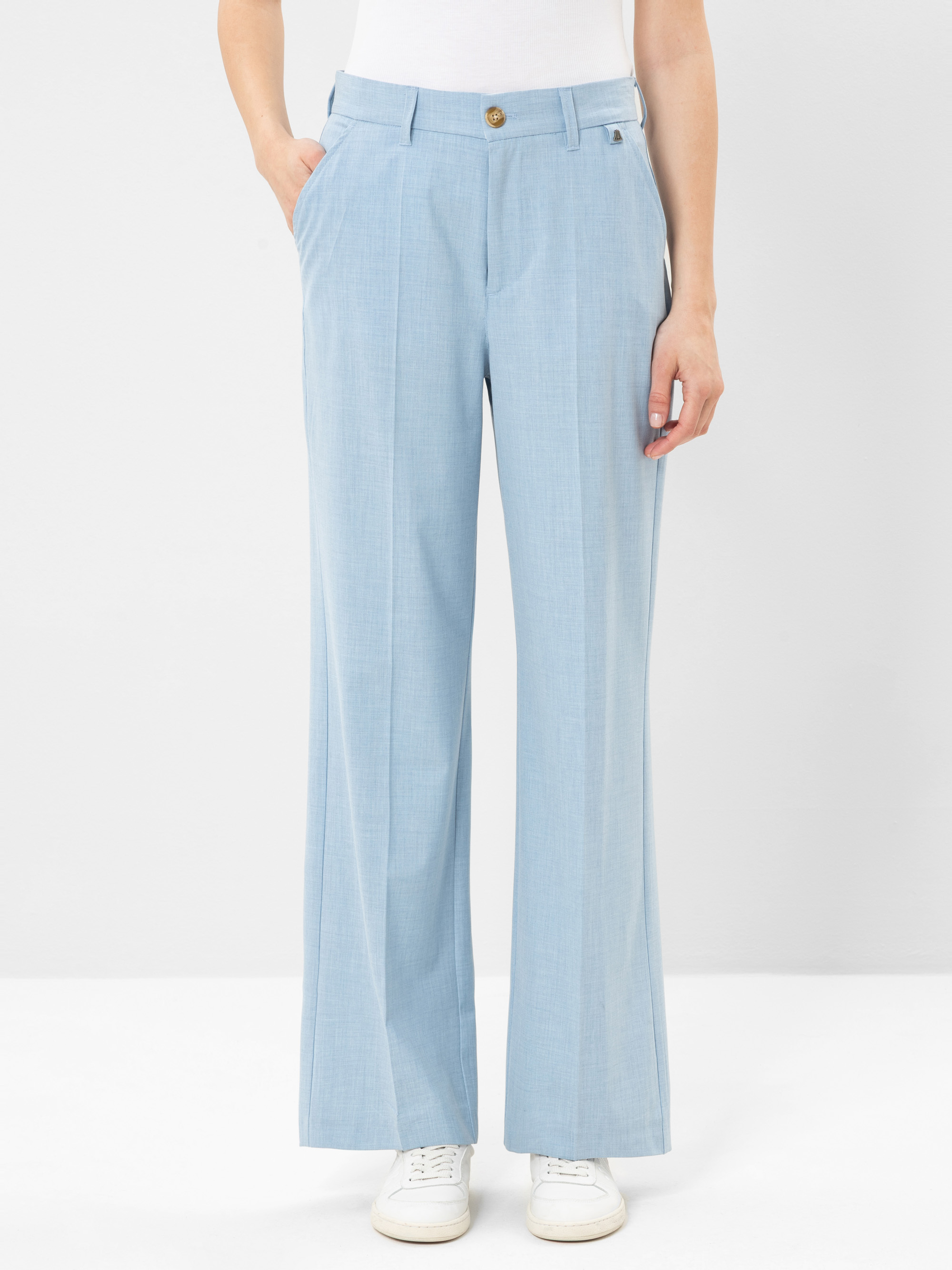 Eine Person, die eine hellblaue Hose mit weitem Bein, ein weißes Oberteil und weiße Turnschuhe trägt, steht mit den Händen in den Taschen vor einem schlichten hellen Hintergrund.