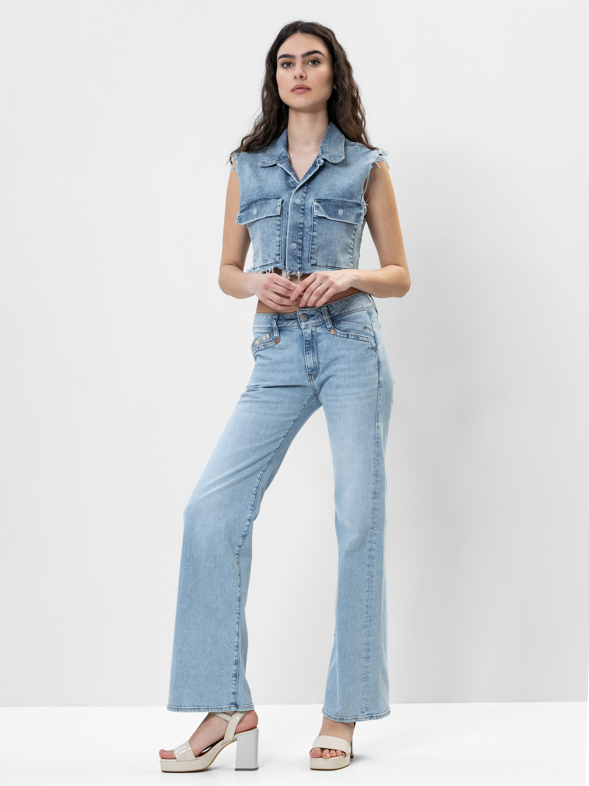 Eine Frau mit langen dunklen Haaren trägt eine ärmellose hellblaue Jeansweste und dazu passende Jeans mit hoher Taille und ausgestelltem Bein. Sie steht selbstbewusst in weißen Plateausandalen vor einem schlichten weißen Hintergrund.