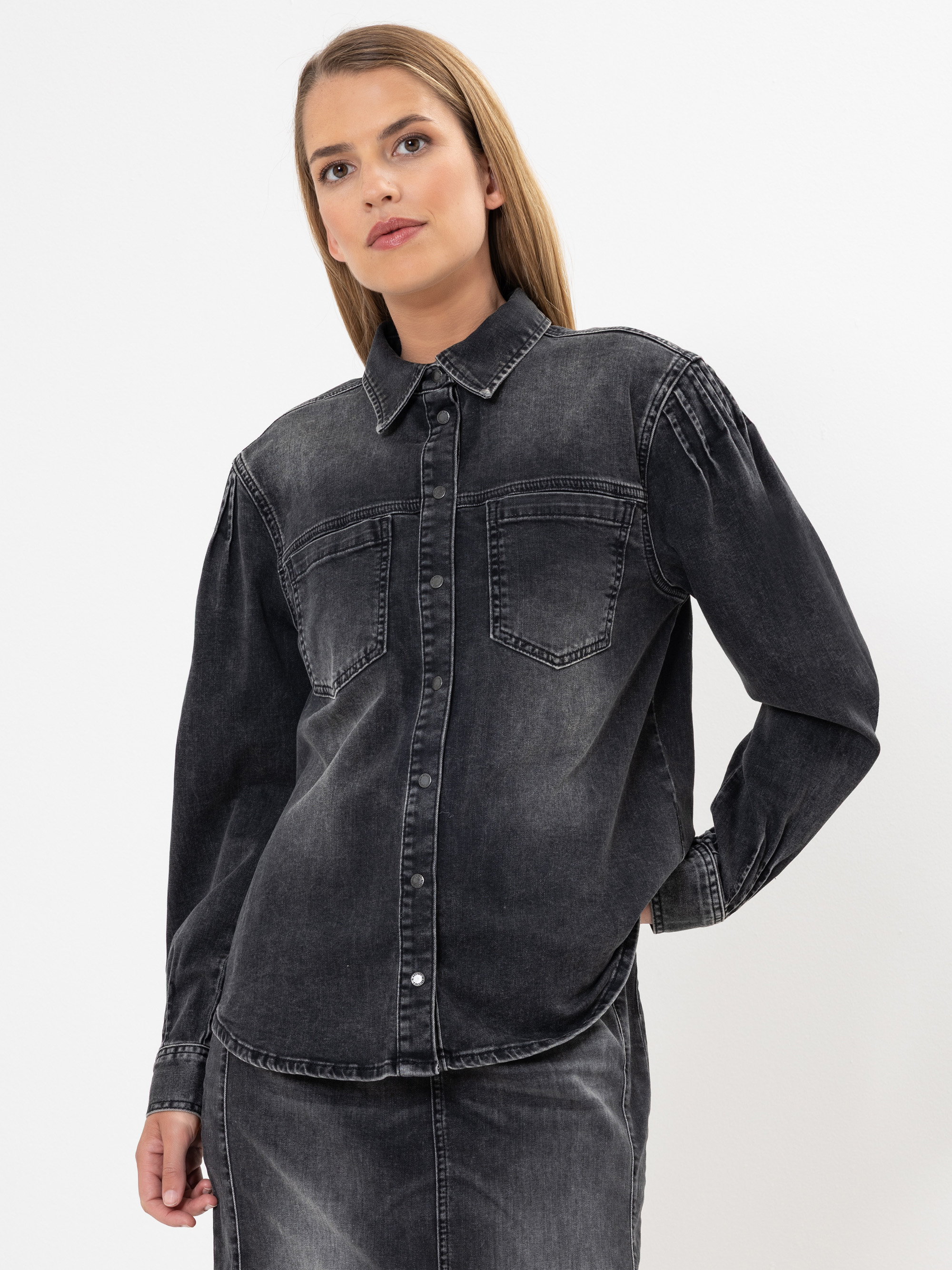 Eine Frau, die ein schwarzes, verblichenes Jeanshemd mit zwei Vordertaschen und einen passenden Rock trägt, steht vor einem schlichten weißen Hintergrund.