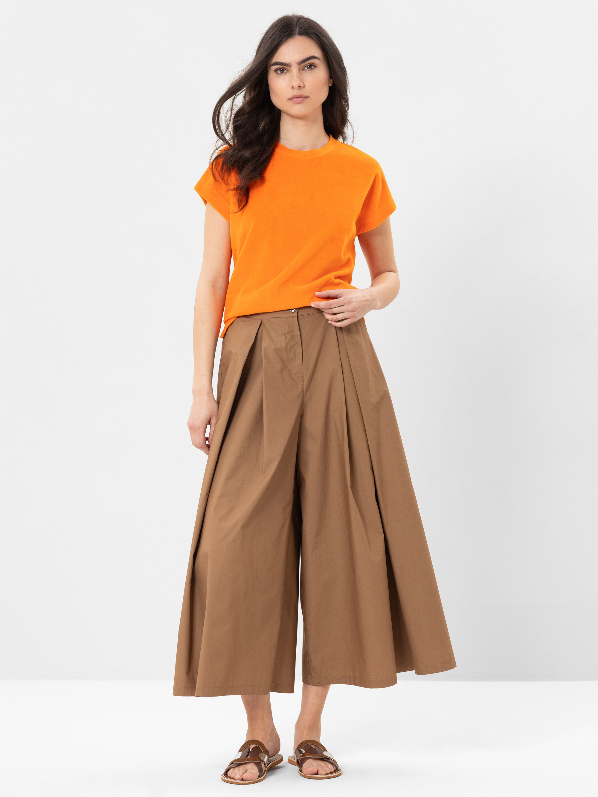 Eine Frau steht vor einem weißen Hintergrund und trägt ein orangefarbenes, kurzärmeliges Oberteil, eine weite, braune, plissierte Culotte-Hose und hellbraune Sandalen. Sie hat langes dunkles Haar und einen neutralen Gesichtsausdruck.