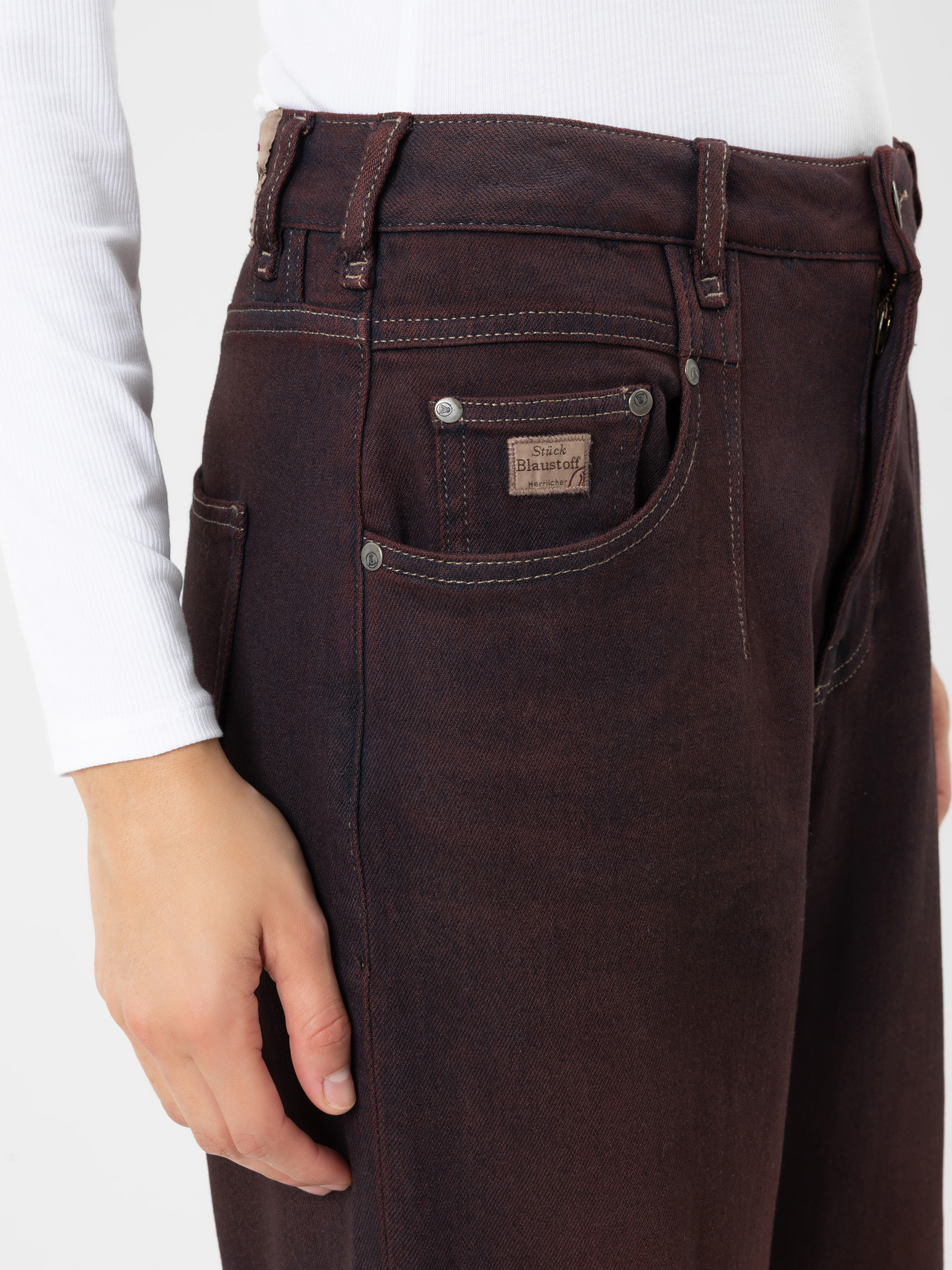 Eine Person, die eine dunkelbraune Jeans mit hoher Taille und sichtbaren Nähten sowie ein weißes, geripptes Langarm-Oberteil trägt. Die Hand ruht an der Seite, und die Jeans weist an der Münztasche ein Markenetikett auf.