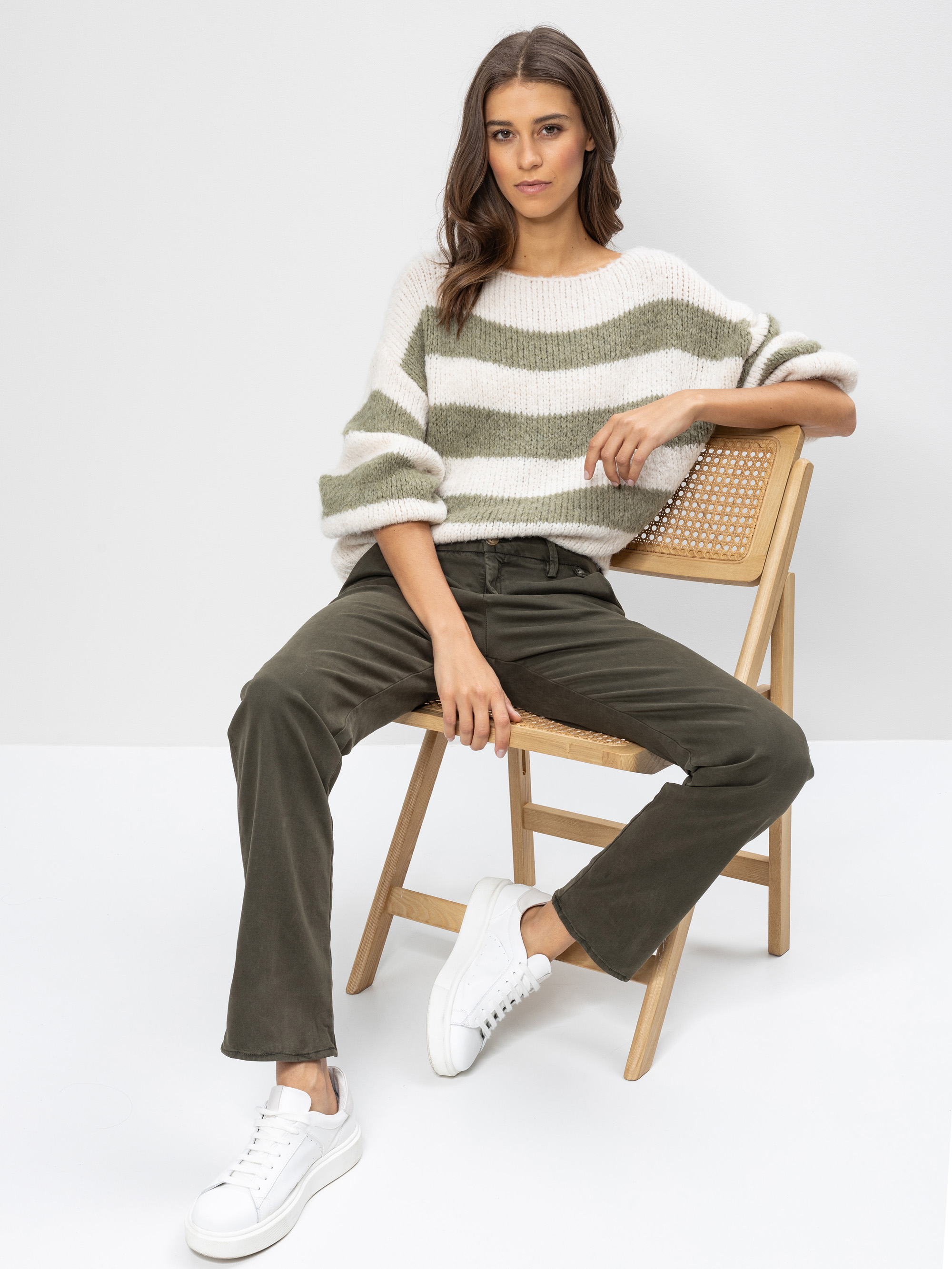 Eine Frau sitzt auf einem Holzstuhl vor einem schlichten Hintergrund und trägt einen übergroßen gestreiften Pullover, eine olivgrüne Hose und weiße Turnschuhe. Sie hat langes braunes Haar und einen entspannten, selbstbewussten Gesichtsausdruck.