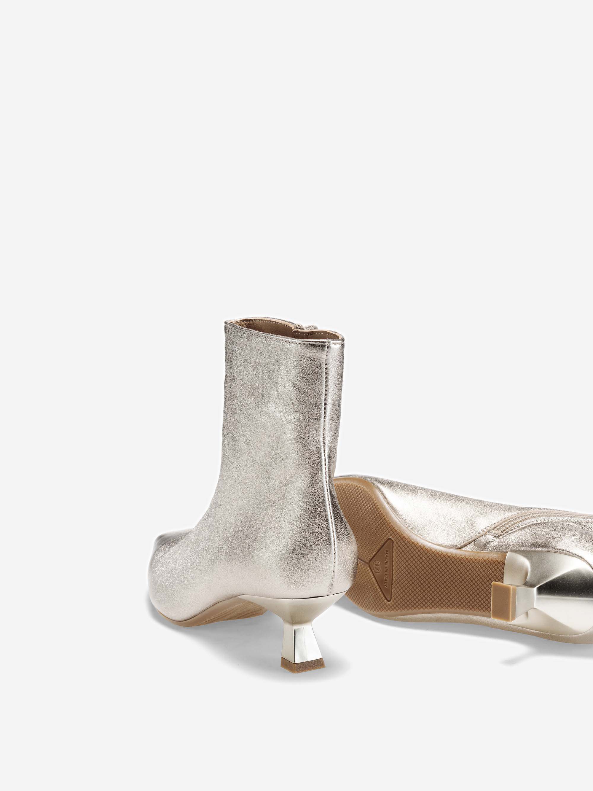 Ein Paar silbern glänzende Stiefeletten mit kurzen, kantigen Absätzen. Ein Stiefel steht aufrecht, während der andere auf der Seite liegt und die Sohle zeigt. Der Hintergrund ist einfarbig weiß.