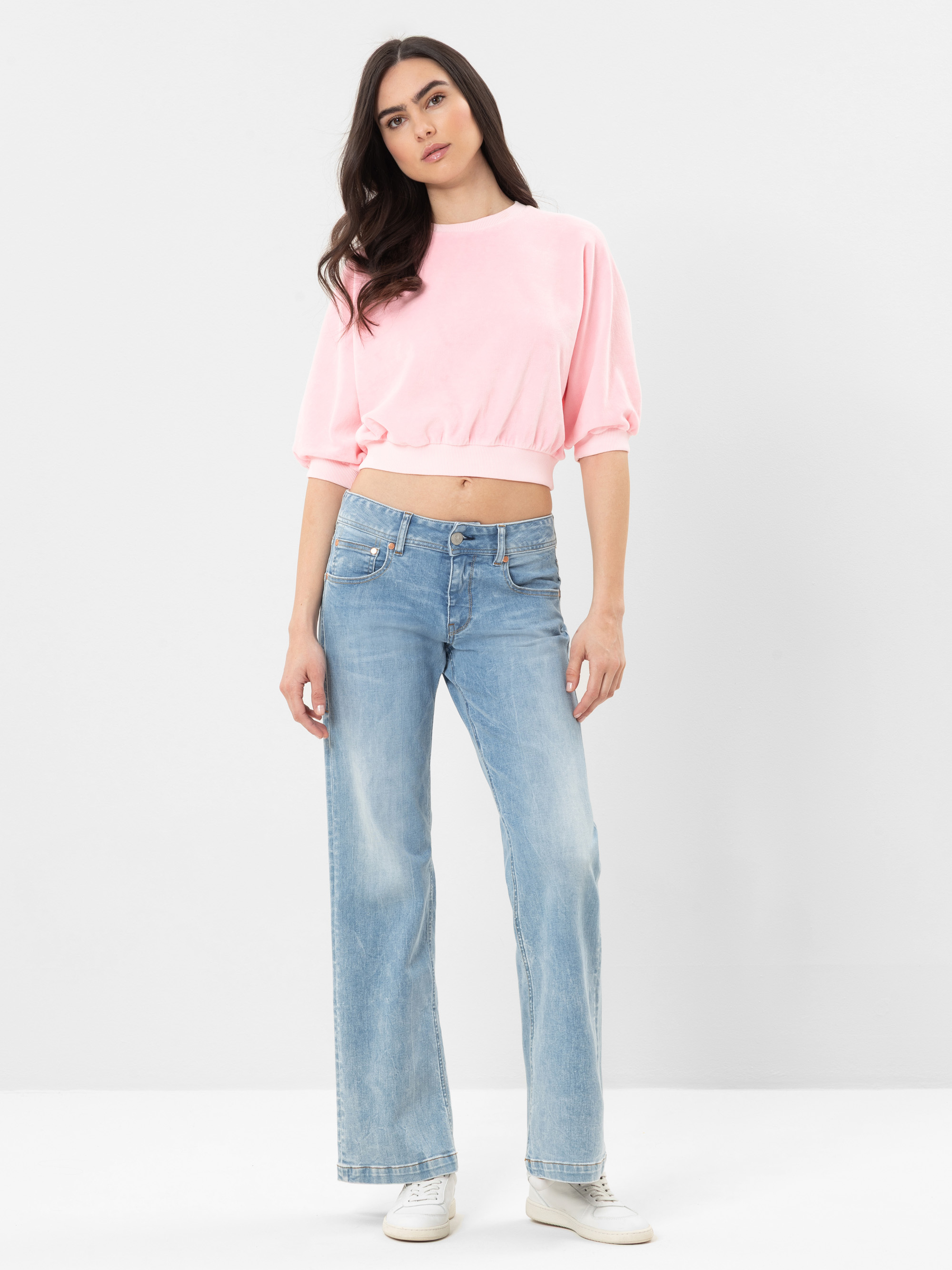 Eine Frau steht vor einem schlichten weißen Hintergrund und trägt ein hellrosa Oberteil, hellblaue Jeans mit weitem Bein und weiße Turnschuhe. Sie hat langes, gewelltes dunkles Haar und einen neutralen Gesichtsausdruck.