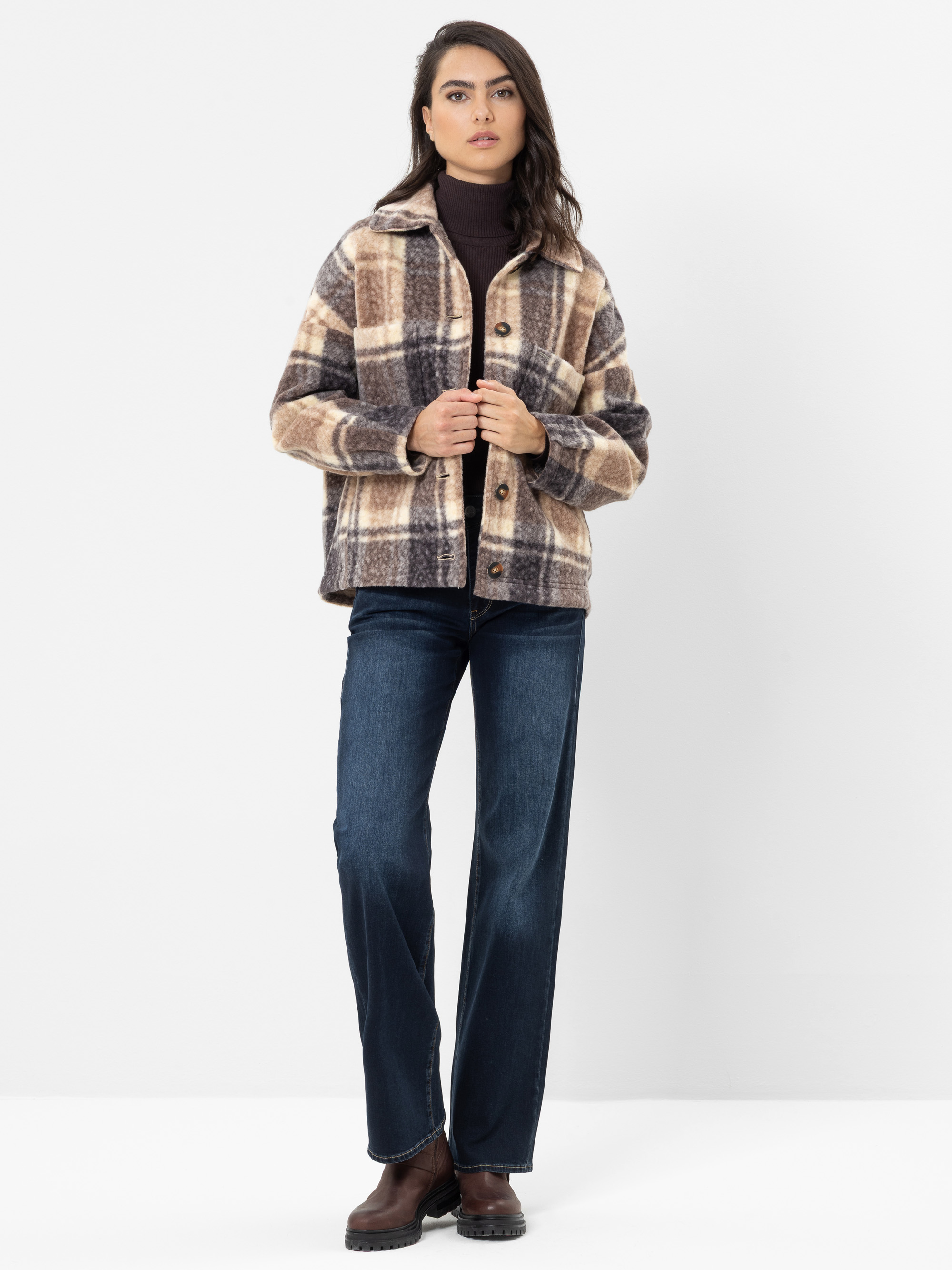 Eine Frau in einer braunen Karojacke, einem dunklen Rollkragenpullover, blauen Jeans und braunen Stiefeln steht vor einem schlichten weißen Hintergrund, blickt nach vorne und zieht mit den Händen ihre Jacke zurecht.