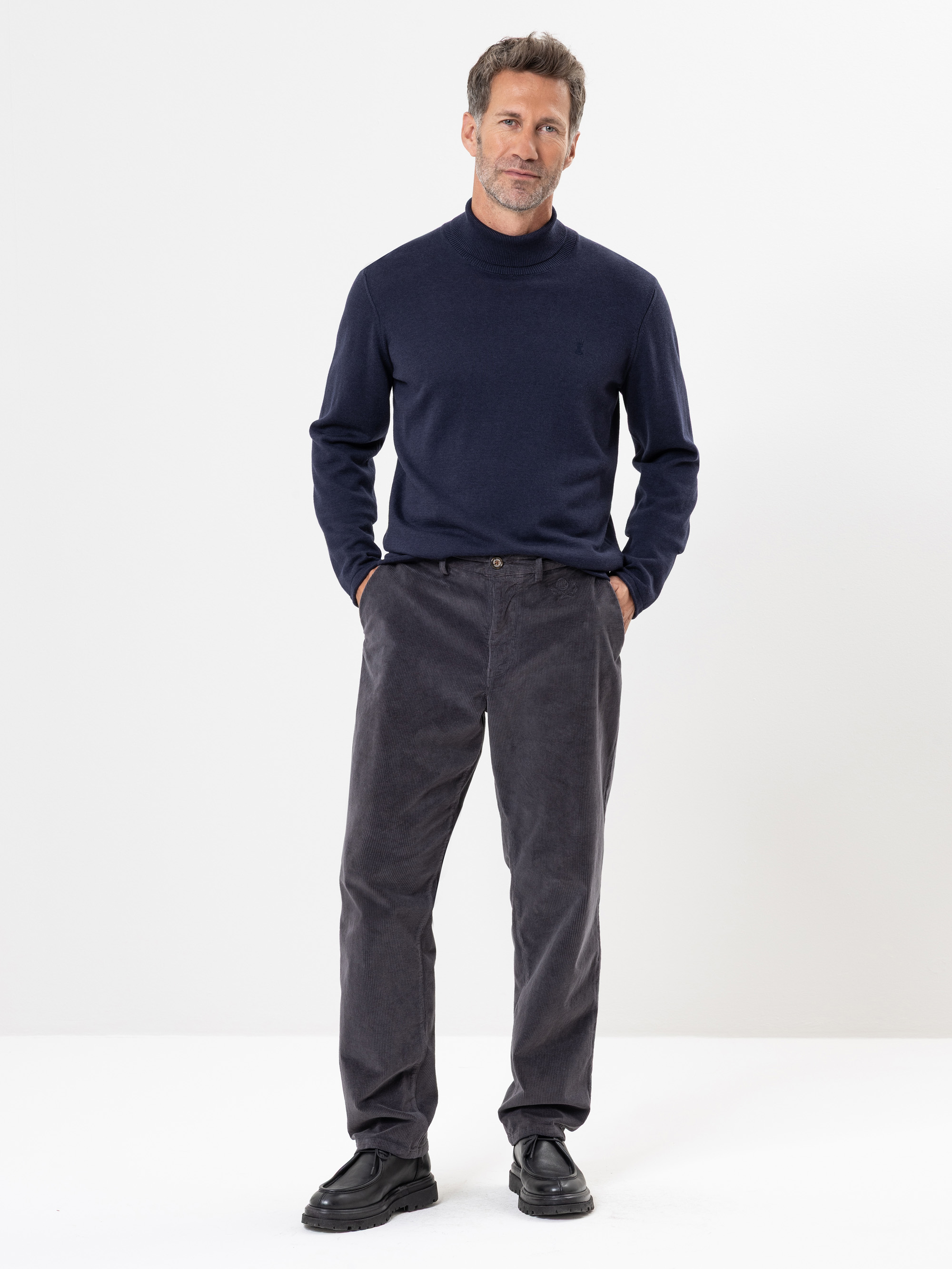 Ein Mann steht vor einem weißen Hintergrund und trägt einen marineblauen Rollkragenpullover, eine graue Kordhose und schwarze Schuhe. Er hat die Hände in den Hosentaschen und blickt mit neutraler Miene nach vorne.