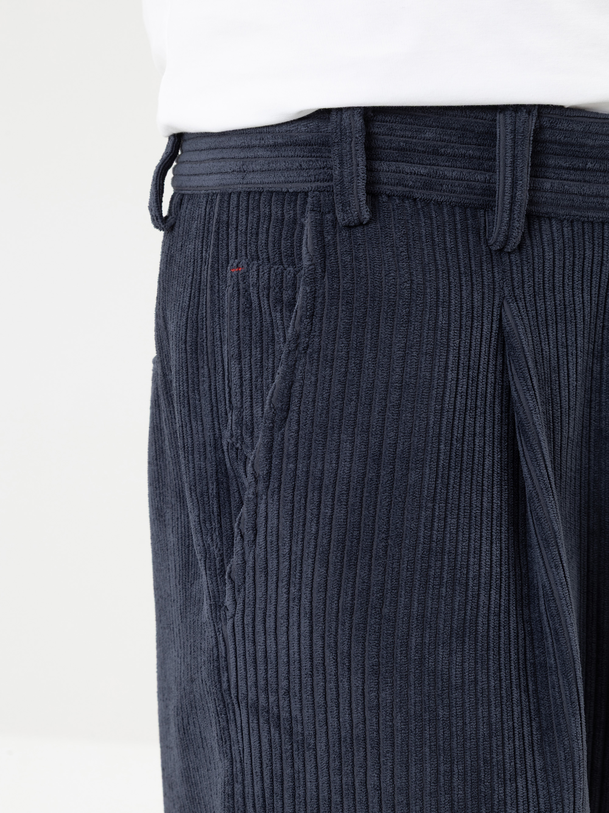Eine Nahaufnahme einer Person, die eine dunkelblaue Cordhose und ein weißes Hemd trägt, wobei der Fokus auf dem Hosenbund, der Tasche und den strukturierten vertikalen Rillen des Stoffes liegt.