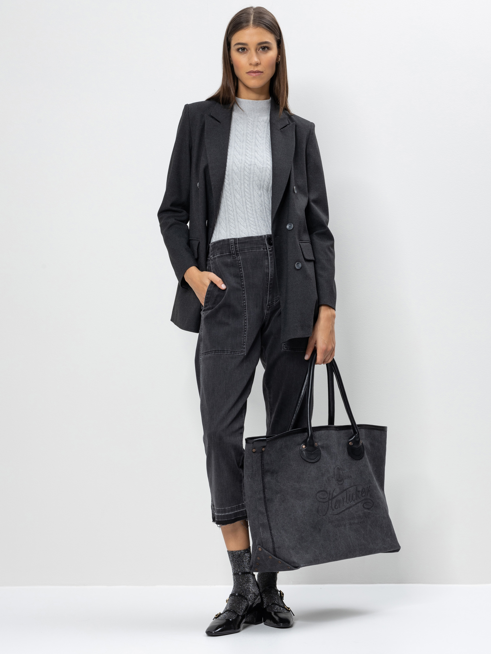 Eine Frau steht vor einem schlichten Hintergrund, trägt einen dunklen Blazer, einen hellgrauen Pullover, dunkle Jeans, schwarze Schuhe und hält eine große dunkle Tragetasche mit der Aufschrift Start Now in der Hand.