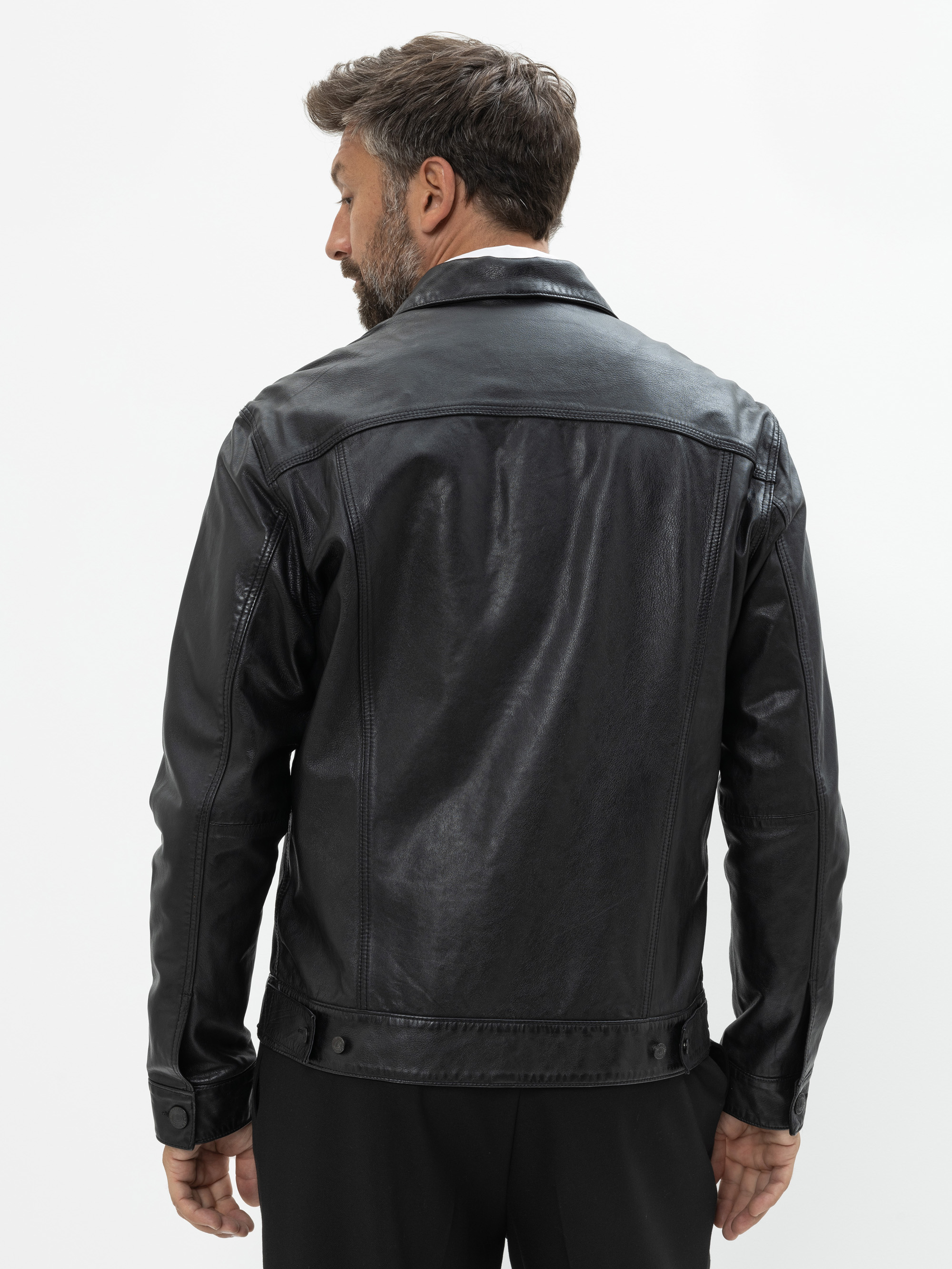 Ein Mann mit kurzen braunen Haaren und einem Bart ist von hinten zu sehen. Er trägt eine schwarze Lederjacke und eine schwarze Hose vor einem schlichten weißen Hintergrund.