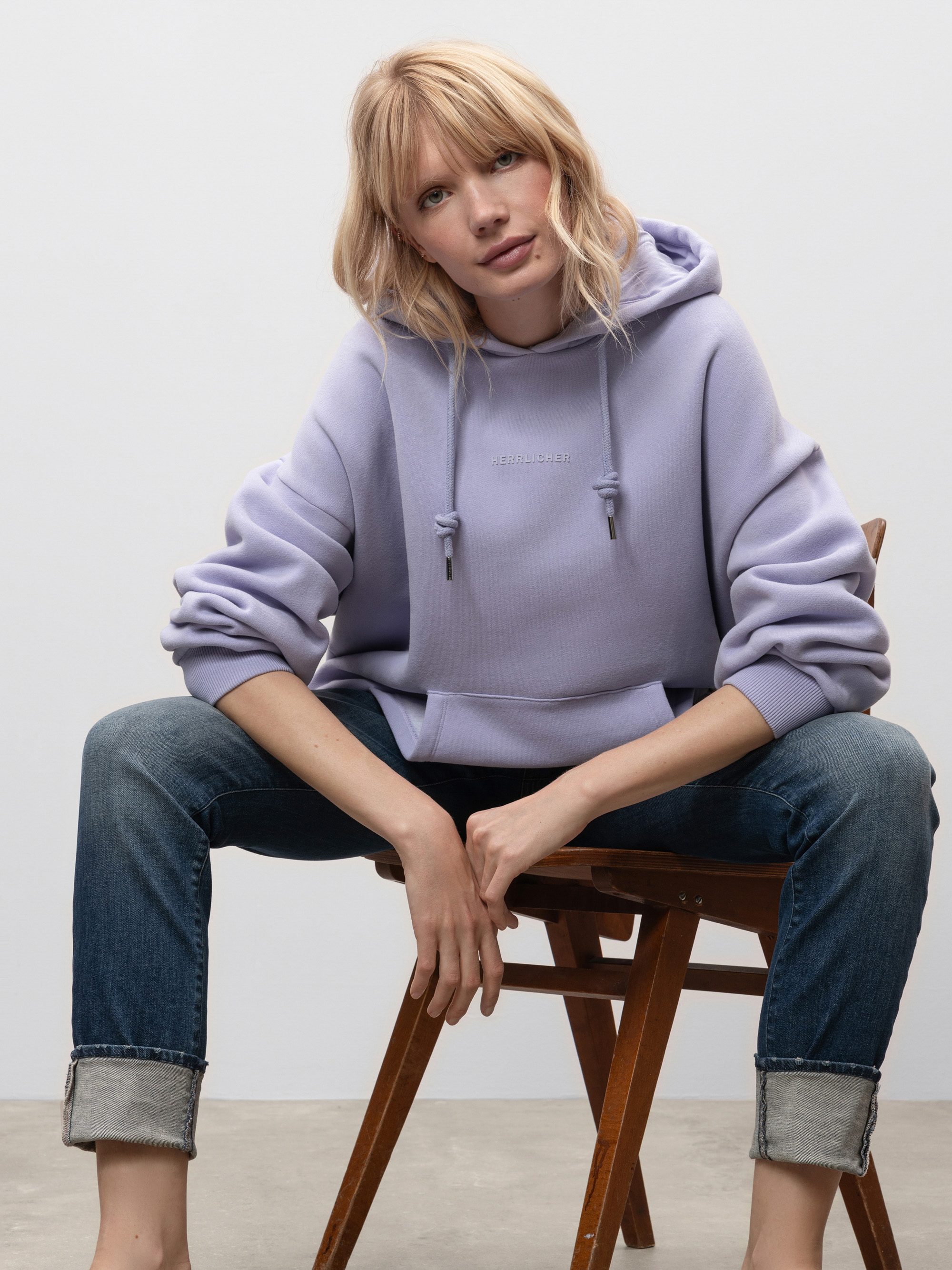 Eine Frau mit blondem Haar, lavendelfarbenem Kapuzenpulli und Jeans mit Manschetten sitzt lässig auf einem Holzstuhl vor einem schlichten hellen Hintergrund und blickt direkt in die Kamera.