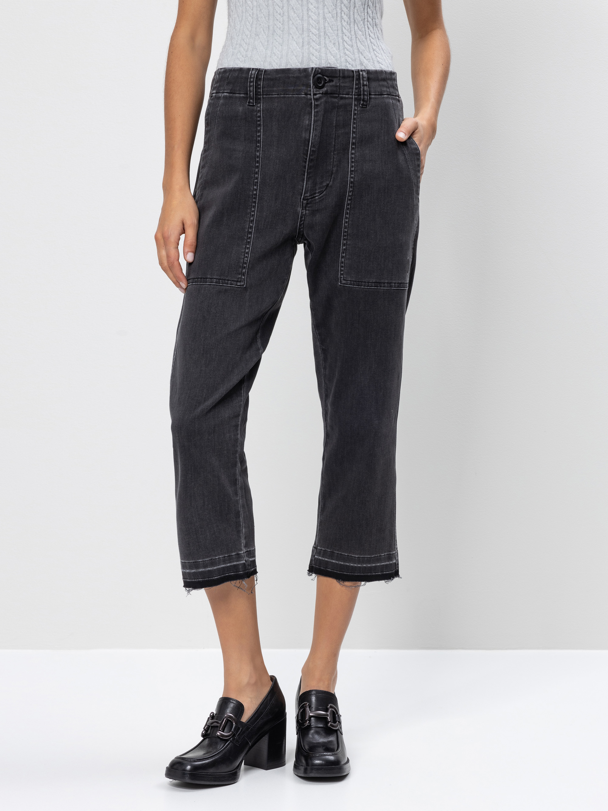 Eine Person trägt eine schwarze Jeans mit aufgesetzten Vordertaschen, einen hellgrauen ärmellosen Pullover und schwarze Halbschuhe mit Absatz vor einem schlichten hellgrauen Hintergrund.