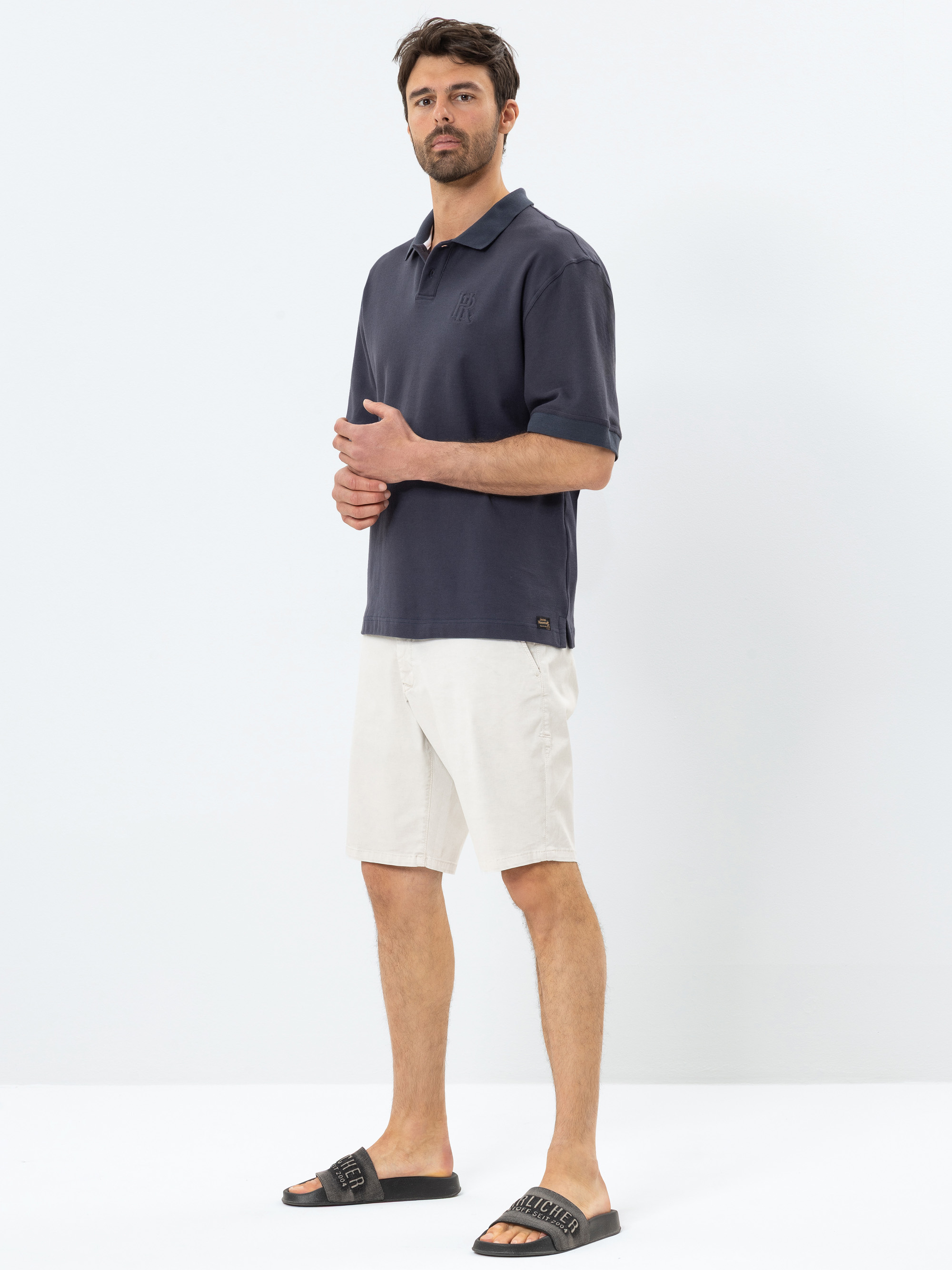 Ein Mann steht vor einem schlichten weißen Hintergrund und trägt ein dunkelblaues Poloshirt, hellbeige Shorts und schwarze Slide-Sandalen. Er blickt nach vorne und hat die Hände locker gefaltet.