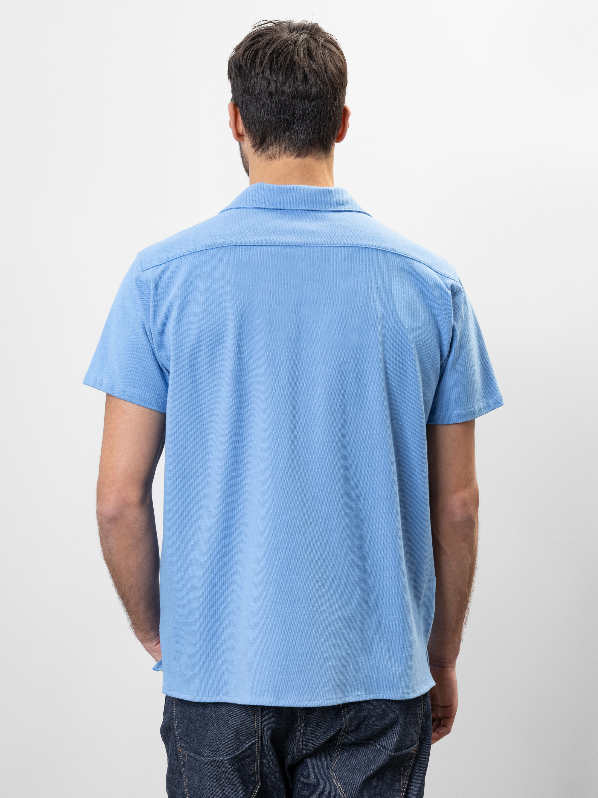 Ein Mann mit kurzen dunklen Haaren steht mit dem Rücken zur Kamera. Er trägt ein hellblaues, kurzärmeliges Poloshirt und dunkle Jeans vor einem schlichten weißen Hintergrund.