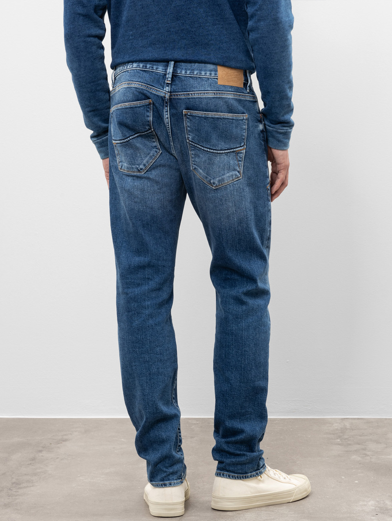 Eine Person steht mit dem Gesicht zur Wand, trägt einen blauen Pullover, eine blaue Jeans mit sichtbaren Taschen und Nähten sowie weiße Turnschuhe und steht vor einer schlichten hellen Wand und einem hellen Boden.