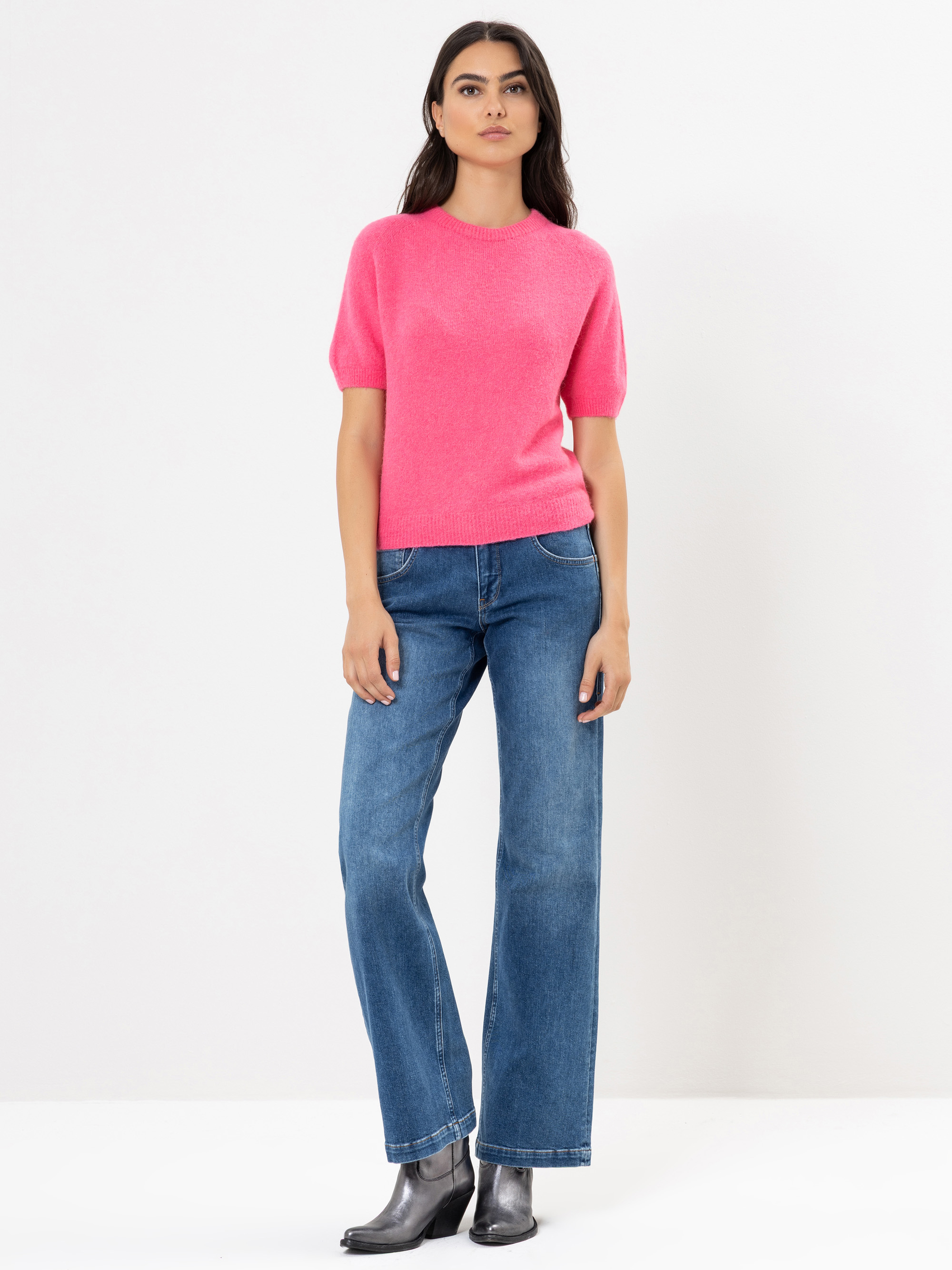 Eine Frau steht vor einem schlichten weißen Hintergrund und trägt einen hellrosa kurzärmeligen Pullover, blaue Jeans und silberne Metallic-Stiefel. Sie hat langes dunkles Haar und einen neutralen Gesichtsausdruck.