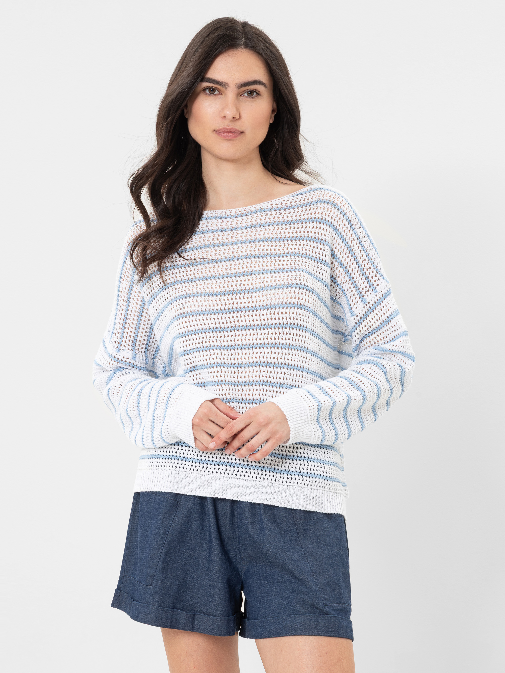 Eine Frau mit langen dunklen Haaren trägt einen lockeren, langärmeligen weißen Pullover mit hellblauen Streifen und dunkelblaue Shorts vor einem schlichten weißen Hintergrund.