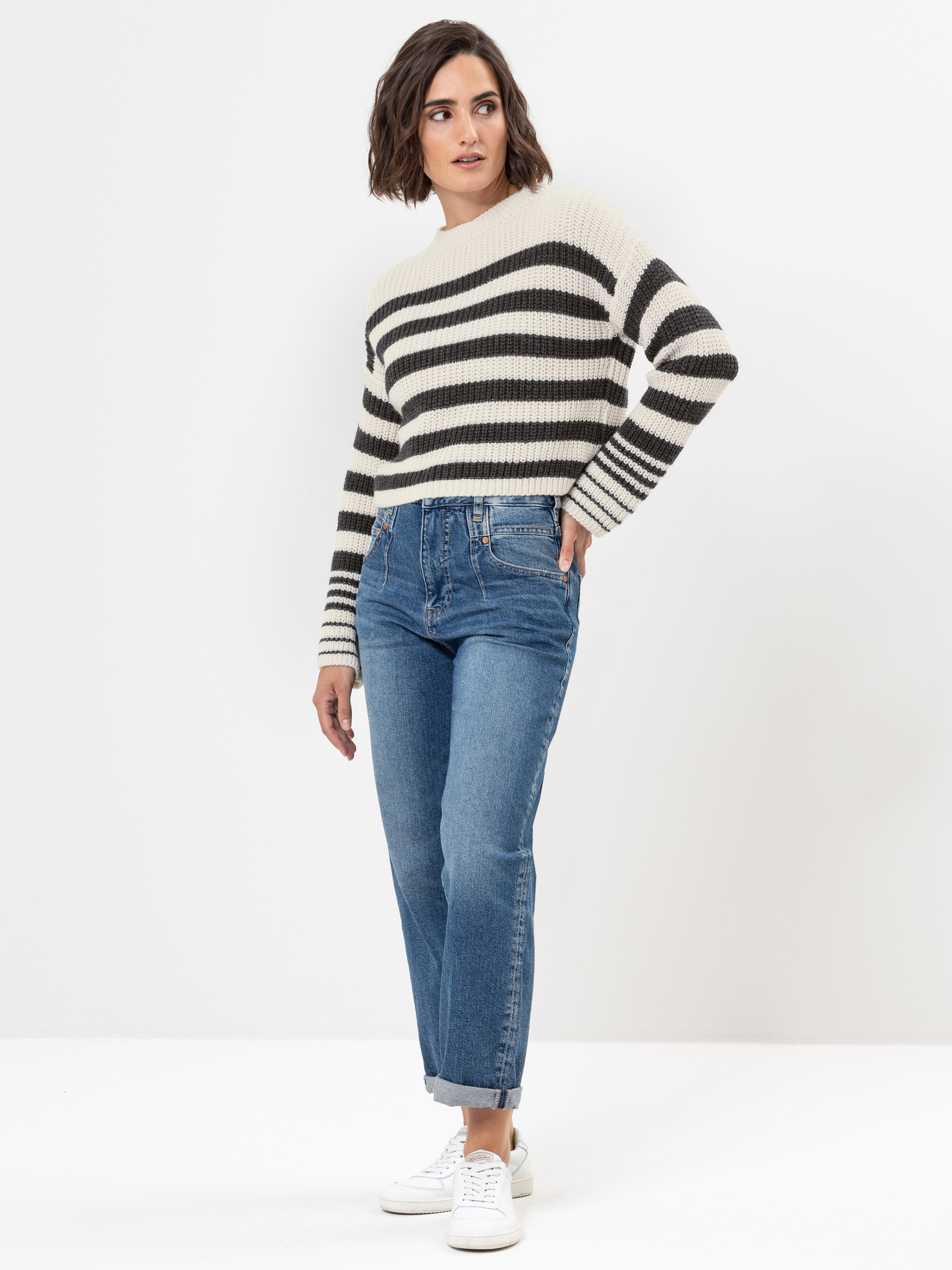 Eine Frau steht vor einem weißen Hintergrund und trägt einen weiß-schwarz gestreiften Pullover, blaue, hochgekrempelte Jeans und weiße Turnschuhe. Sie hat kurzes braunes Haar und posiert lässig mit einer Hand in der Tasche.