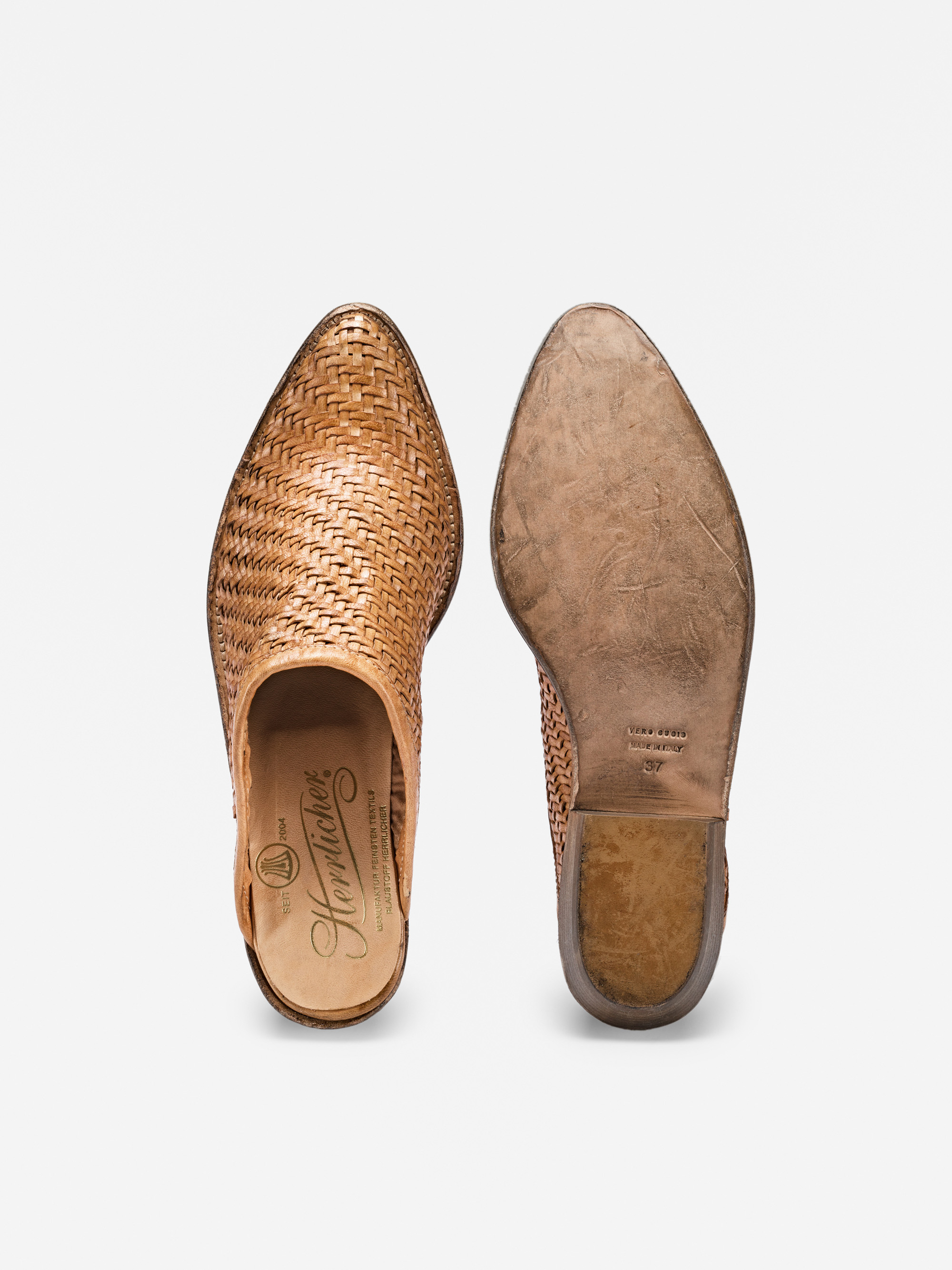 Ein Paar hellbraune Slipper aus gewebtem Leder; einer davon ist von oben abgebildet und zeigt sein kompliziertes Geflecht und die gebrandete Innensohle, während der andere von unten gezeigt wird und eine abgenutzte Ledersohle aufweist.