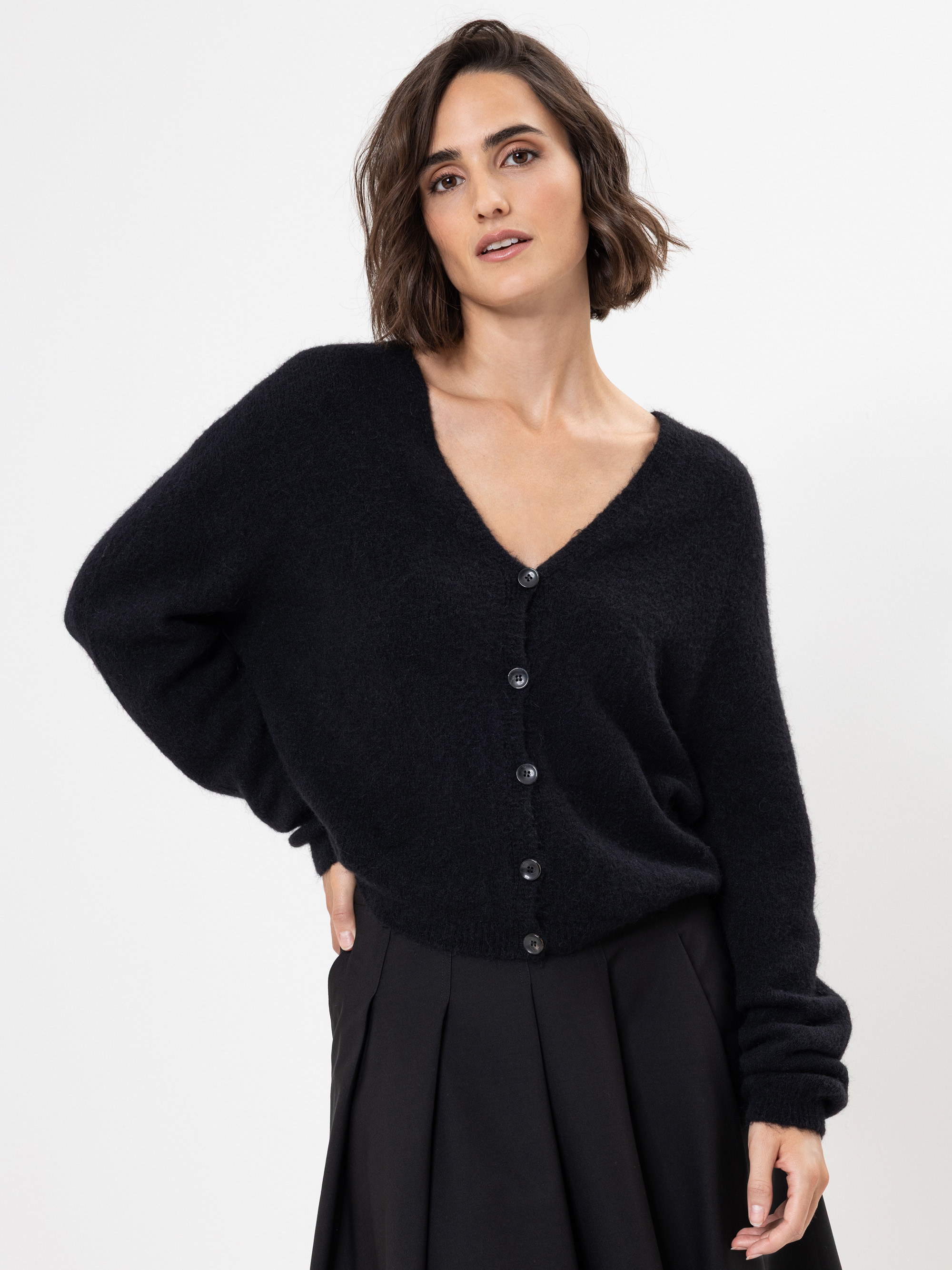 Eine Frau mit einer kurzen braunen Bobfrisur, die einen schwarzen Pullover mit V-Ausschnitt und einen schwarzen Faltenrock trägt, steht vor einem schlichten weißen Hintergrund. Ihre linke Hand ruht auf ihrer Hüfte und ihr rechter Arm ist entspannt.