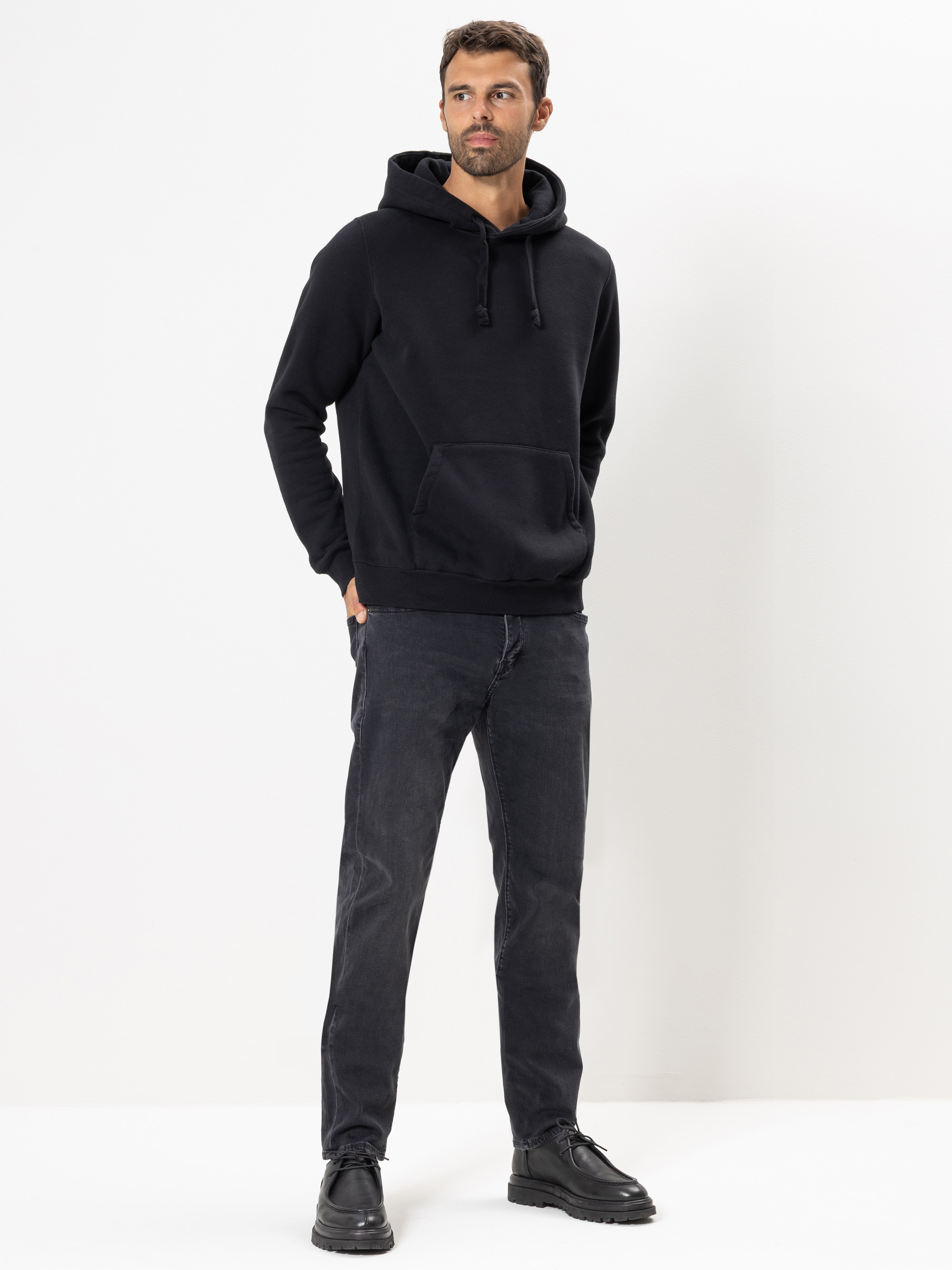 Ein Mann steht vor einem weißen Hintergrund, trägt einen schwarzen Kapuzenpullover, dunkle Jeans und schwarze Schuhe, hat die Hände in den Taschen und schaut leicht zur Seite.