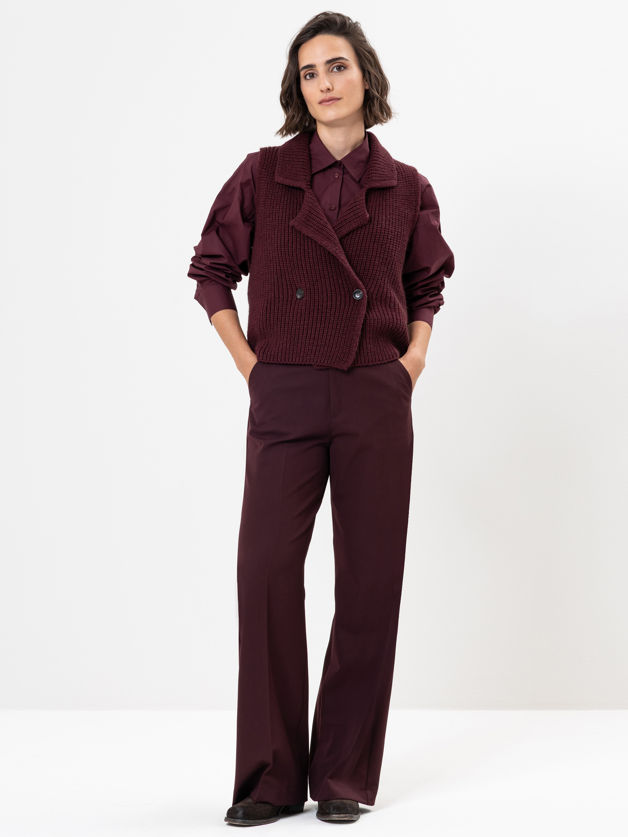 Eine Frau steht vor einem weißen Hintergrund und trägt ein kastanienbraunes Button-up-Hemd, eine passende Hose mit weitem Bein und eine gestrickte kastanienbraune Weste. Ihre Hände stecken in den Taschen und sie hat schulterlanges dunkles Haar.
