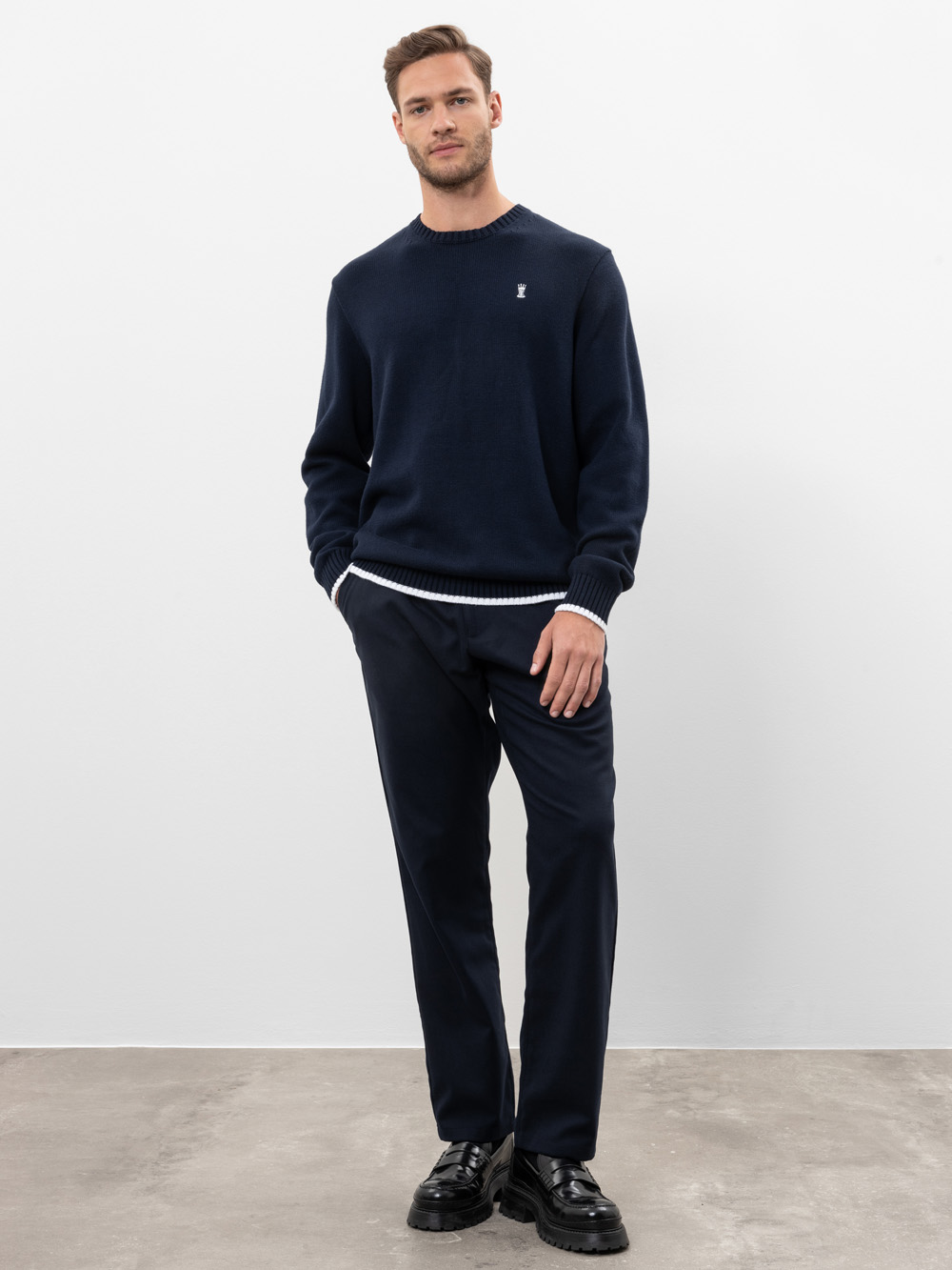 Ein Mann steht vor einer schlichten weißen Wand und trägt einen marineblauen Pullover, eine passende marineblaue Hose und schwarze Anzugschuhe. Er hat eine Hand in der Tasche und schaut mit einem leichten Lächeln in die Kamera.