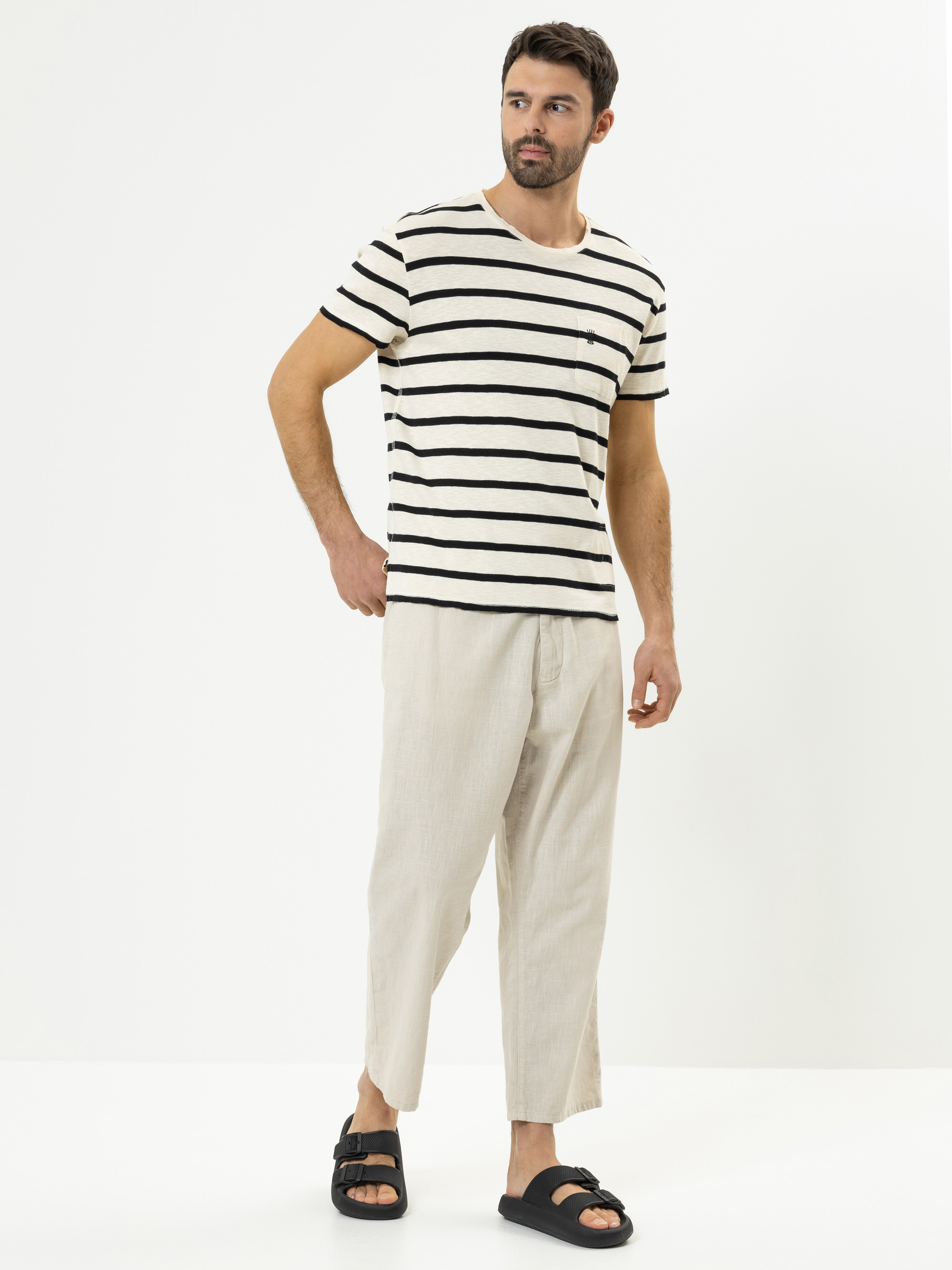 Ein Mann steht vor einem schlichten weißen Hintergrund und trägt ein beige-schwarz gestreiftes T-Shirt, eine weite beigefarbene Hose und schwarze Sandalen. Er hat dunkles Haar und einen kurzen Bart und blickt leicht zur Seite.