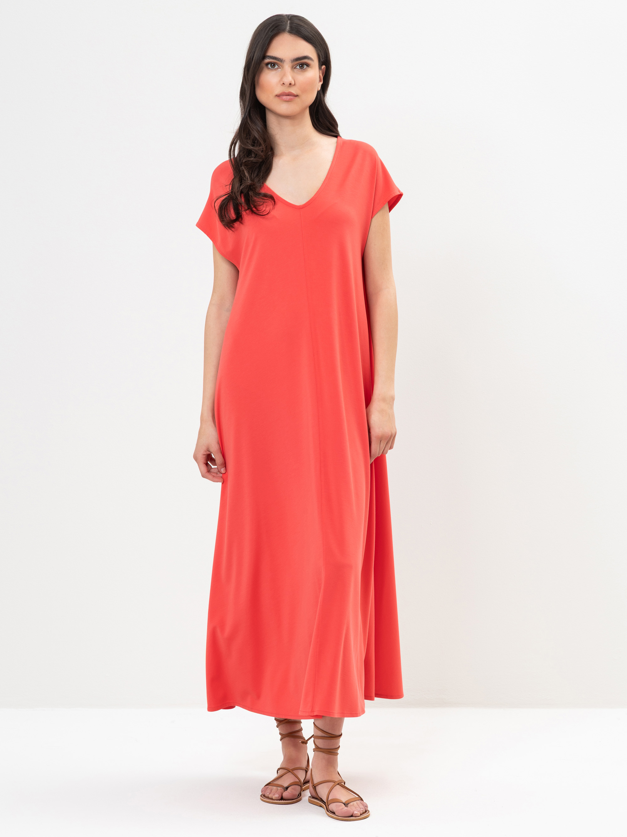 Eine Frau mit langen dunklen Haaren, die ein locker sitzendes, kurzärmeliges, korallenrotes Maxikleid und braune Riemchensandalen trägt, steht vor einem schlichten weißen Hintergrund.