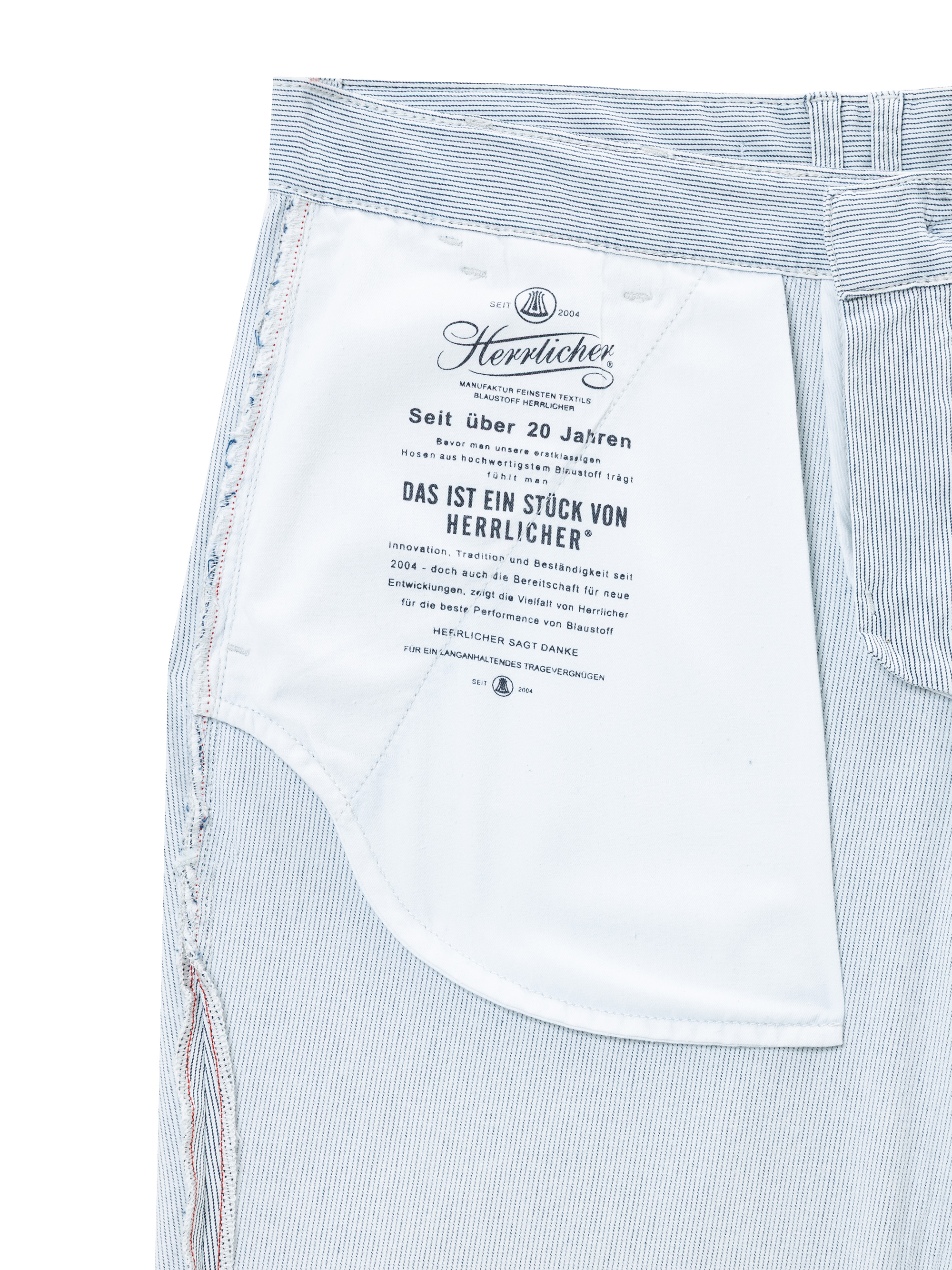 Nahaufnahme der Innentasche einer hellblau-weiß gestreiften Hose, die ein weißes Stoffetikett mit deutschem Text und dem schwarz gedruckten Herrlicher-Logo zeigt.