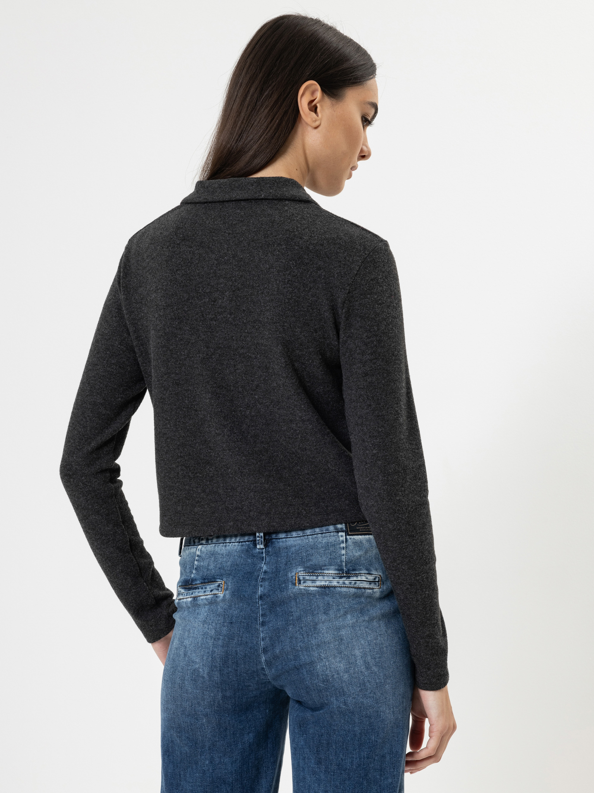 Eine Frau mit langen dunklen Haaren ist von hinten zu sehen. Sie trägt ein dunkelgraues langärmeliges Oberteil und blaue Jeans vor einem schlichten weißen Hintergrund.