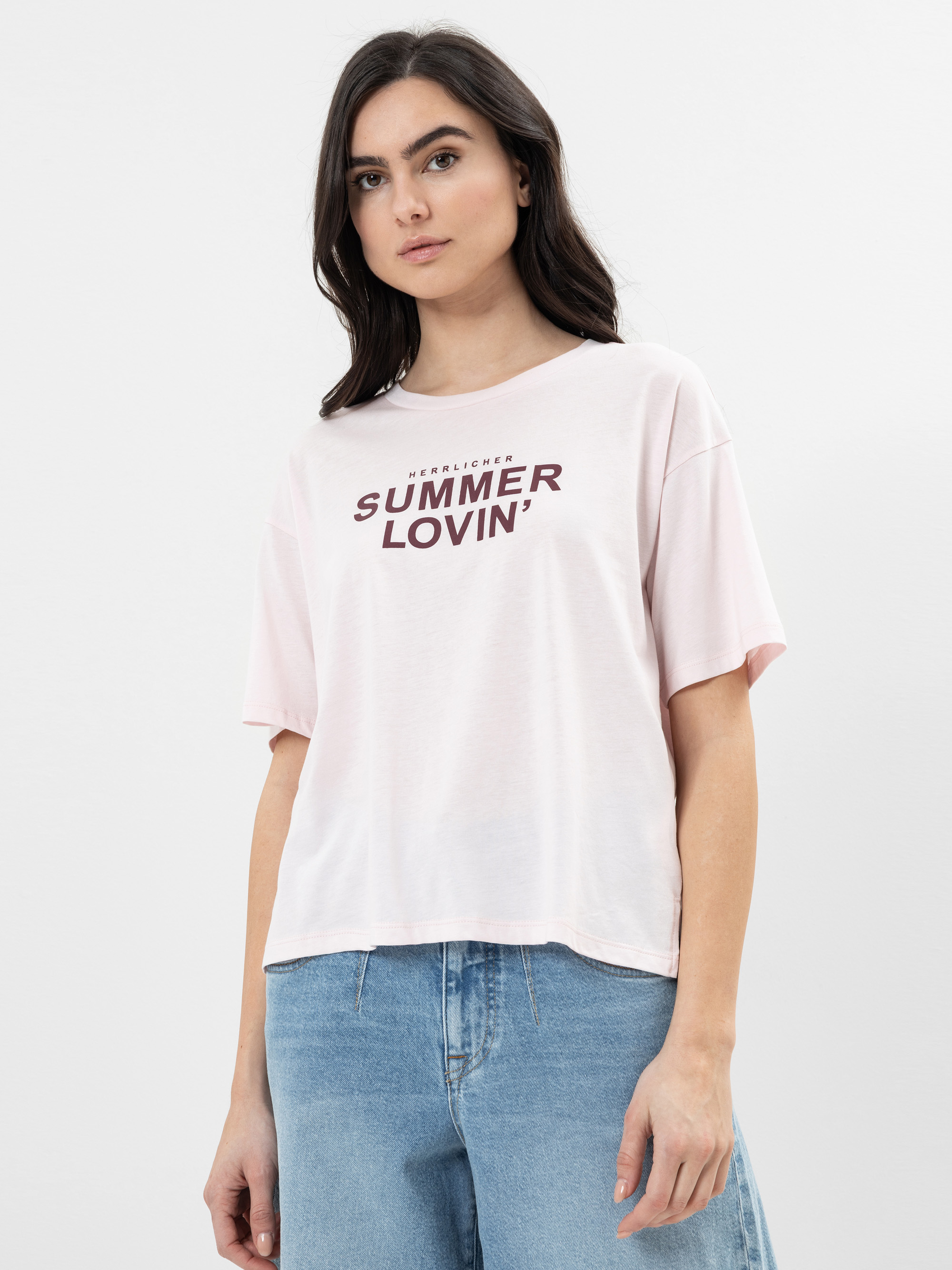 Eine Frau mit langen dunklen Haaren trägt ein hellrosa T-Shirt mit dem Aufdruck "Summer Lovin'" in fetten Buchstaben auf der Vorderseite und hellblaue Jeans, die vor einem schlichten weißen Hintergrund stehen.