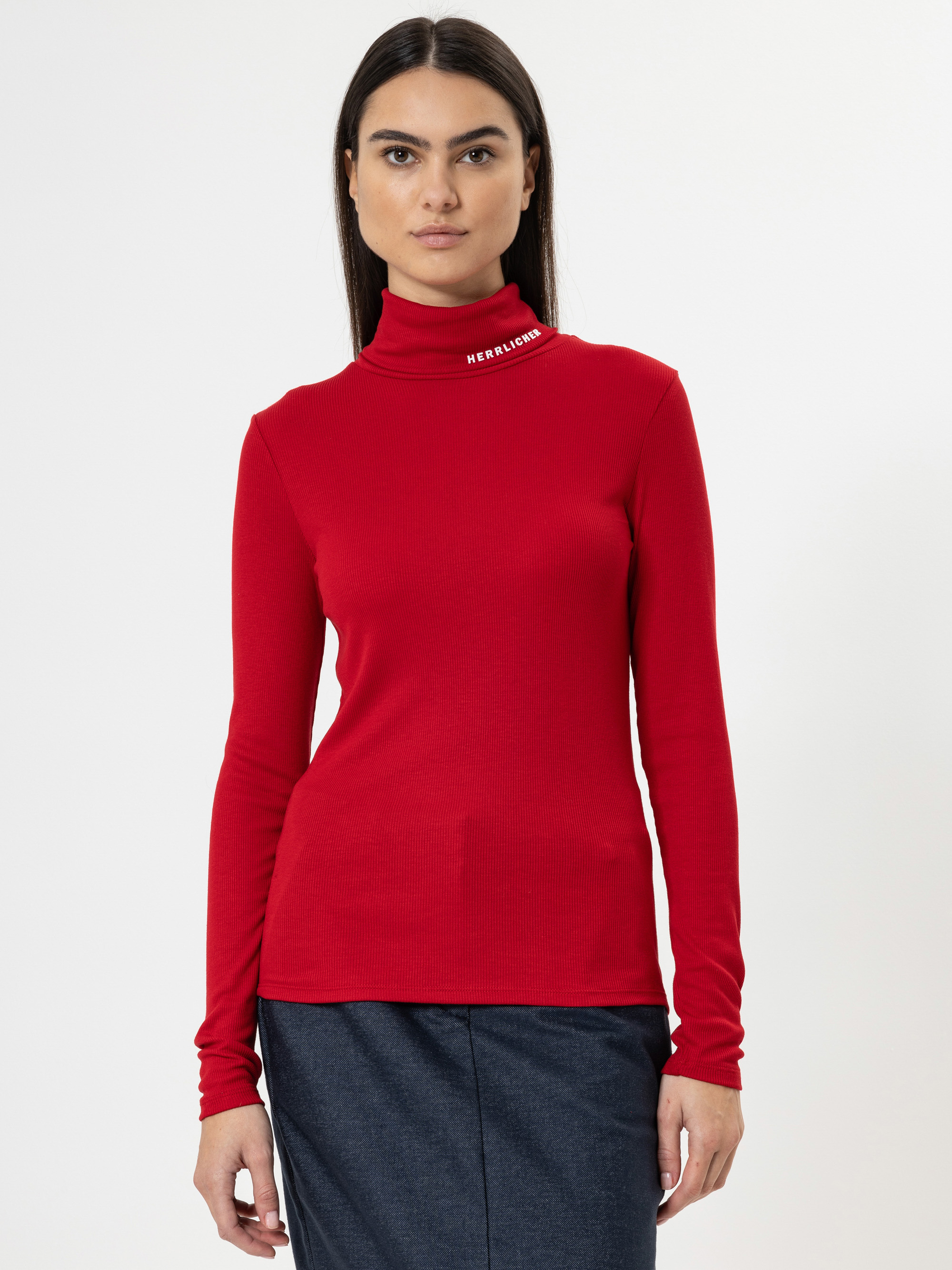 Eine Frau, die einen roten, langärmeligen Rollkragenpullover mit weißer Aufschrift am Kragen und einen dunkelblauen Rock trägt, steht vor einem schlichten weißen Hintergrund und blickt mit neutralem Blick direkt in die Kamera.