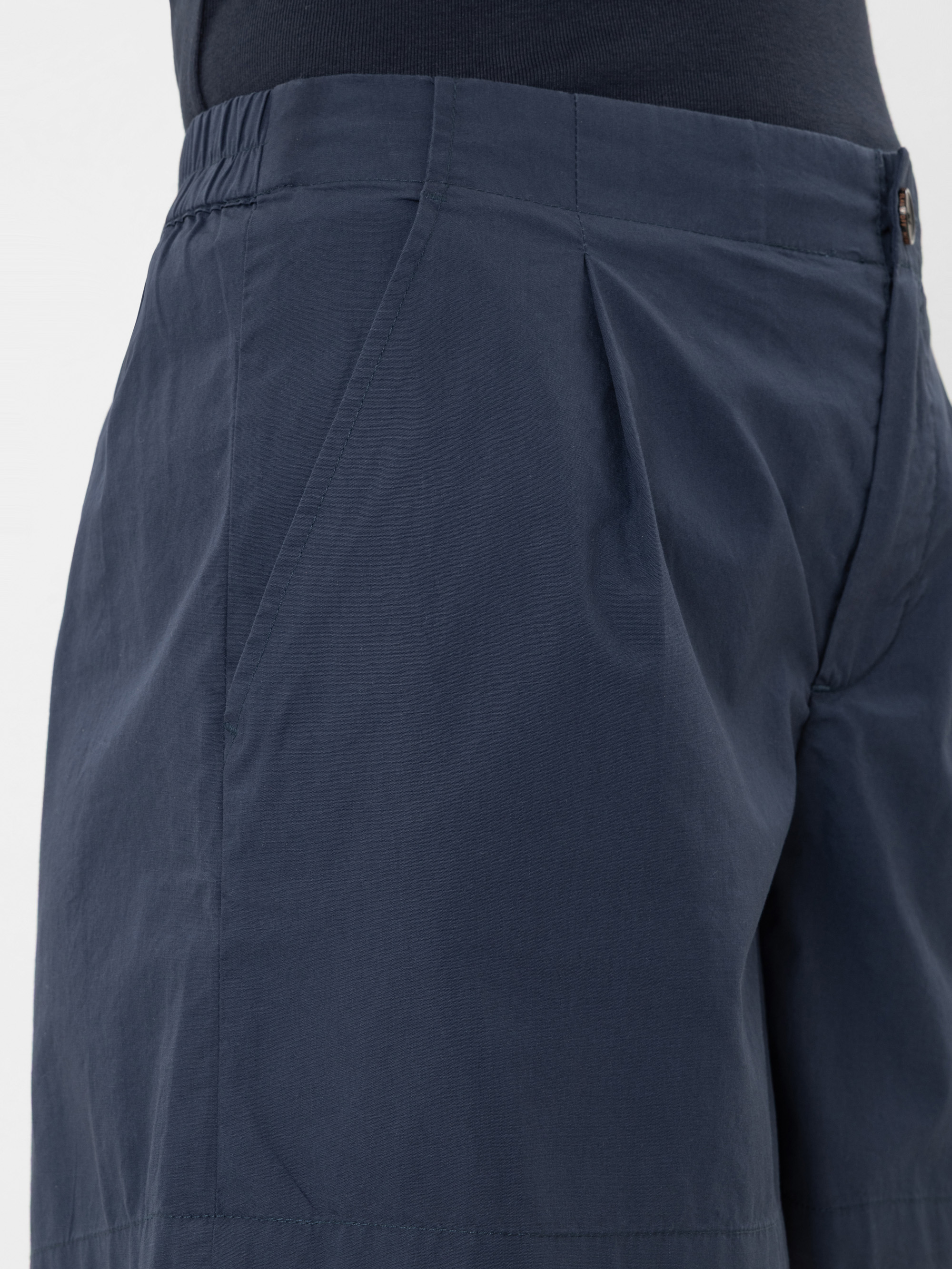 Nahaufnahme einer Person, die dunkelblaue, plissierte Shorts mit Seitentaschen trägt, die den Bund und den Oberschenkelbereich zeigen. Die Shorts scheinen aus leichtem Stoff gefertigt zu sein.