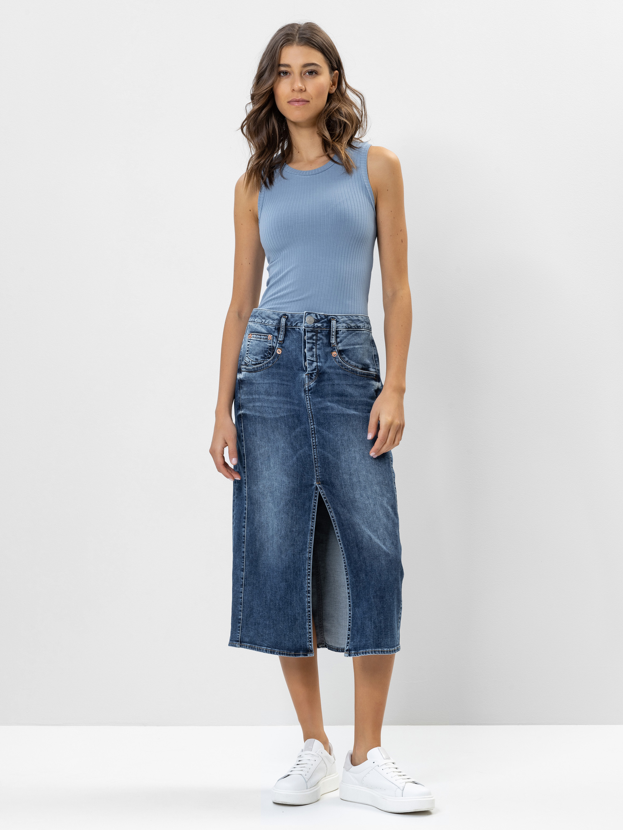 Eine Frau steht vor einem schlichten weißen Hintergrund und trägt ein ärmelloses hellblaues geripptes Oberteil, einen hochtaillierten blauen Jeans-Midirock mit einem Schlitz vorne und weiße Turnschuhe.