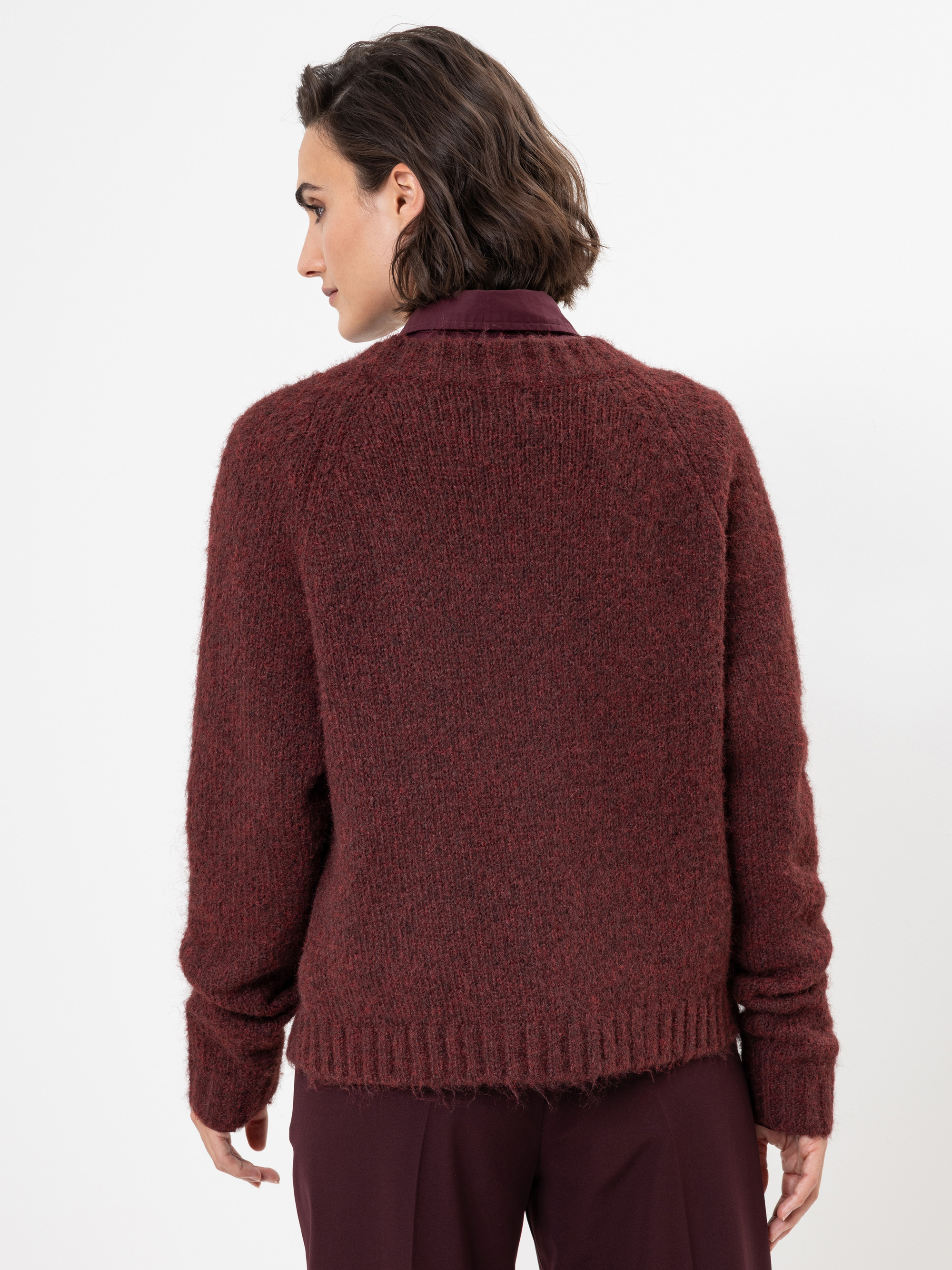 Eine Person mit kurzen braunen Haaren wird von hinten gezeigt, die einen dicken kastanienbraunen Pullover über einem Hemd mit Kragen und eine passende kastanienbraune Hose trägt und vor einem schlichten weißen Hintergrund steht.