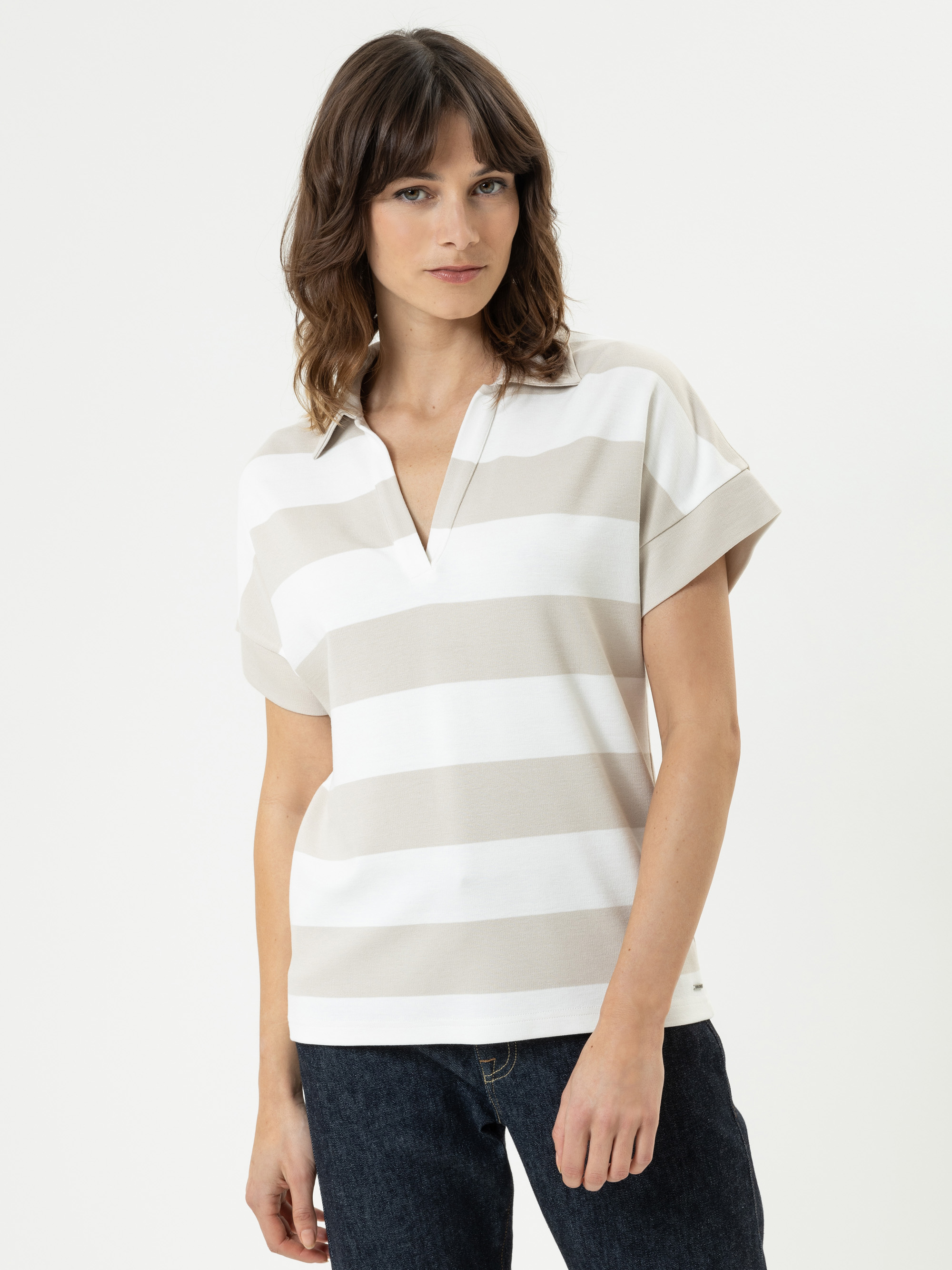 Eine Frau mit gewellten braunen Haaren trägt ein kurzärmeliges, beige-weiß gestreiftes Polohemd und dunkle Jeans und steht vor einem schlichten weißen Hintergrund.