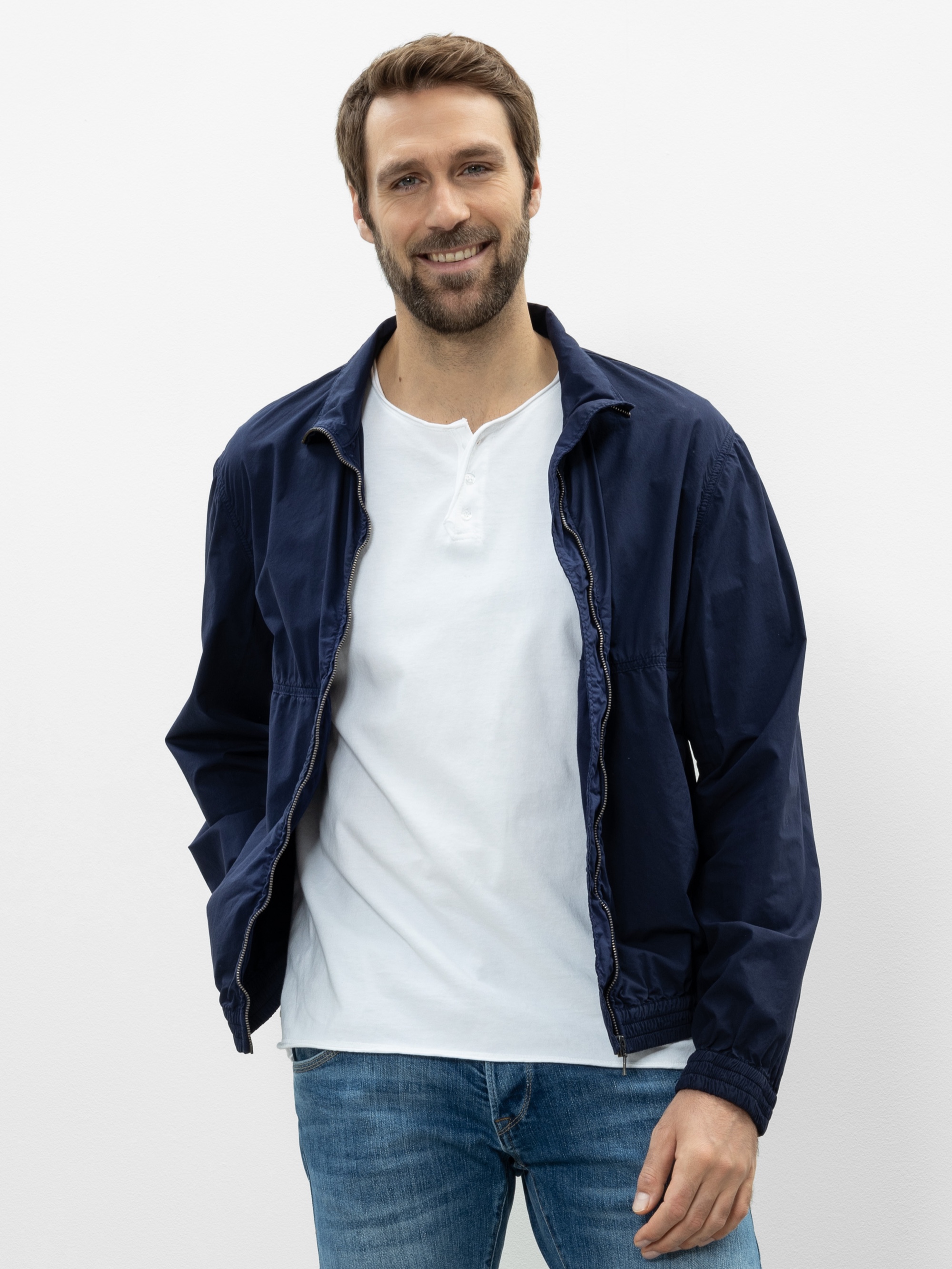 Ein Mann mit kurzen braunen Haaren und einem Bart lächelt. Er trägt eine marineblaue Jacke über einem weißen Hemd und blaue Jeans, steht vor einem schlichten weißen Hintergrund und hat die Hände in den Jackentaschen.