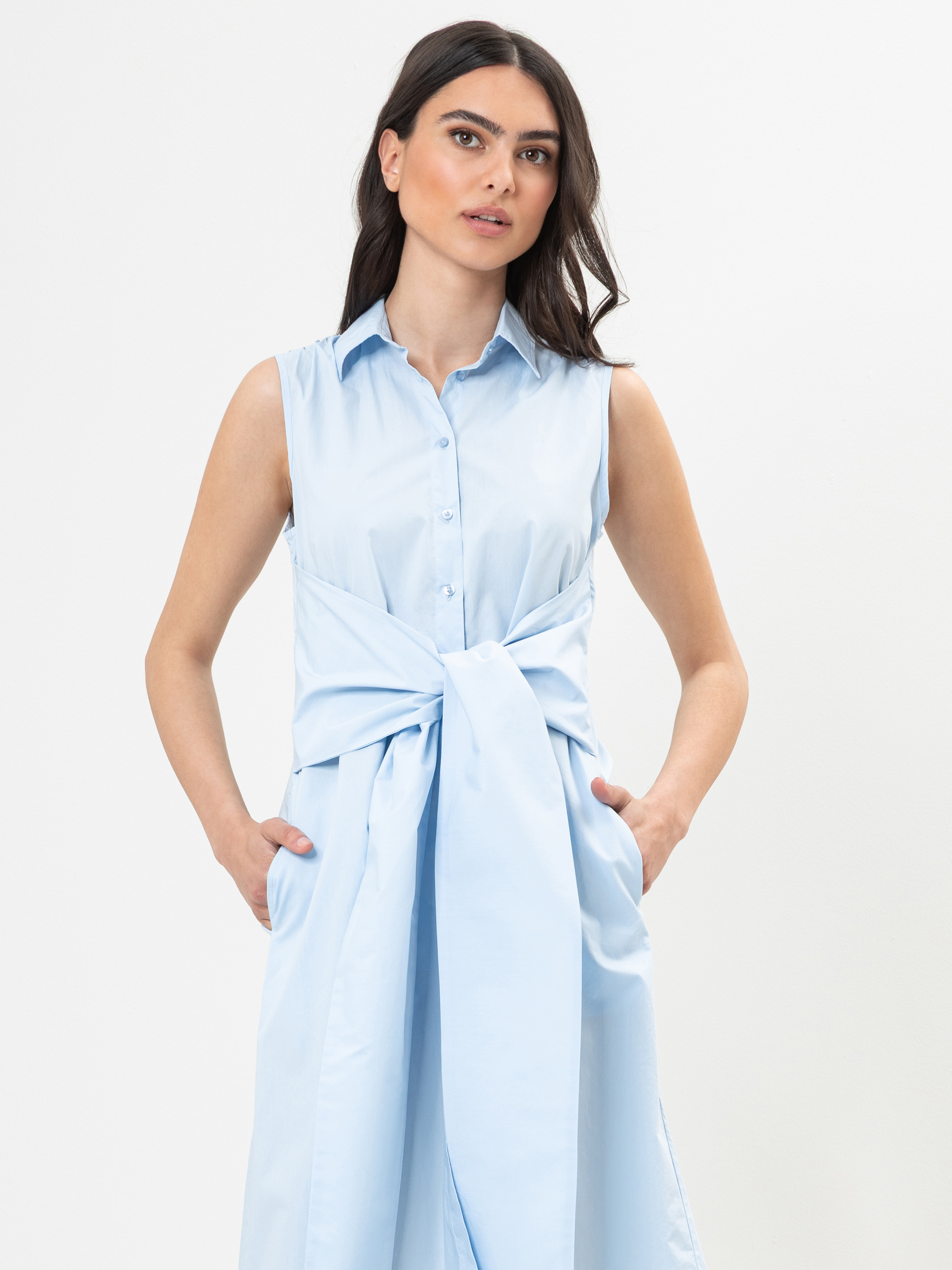Eine Frau mit langen dunklen Haaren, die ein ärmelloses, hellblaues Kleid mit Kragen und einer breiten Krawatte trägt, steht mit den Händen in den Taschen vor einem schlichten weißen Hintergrund.