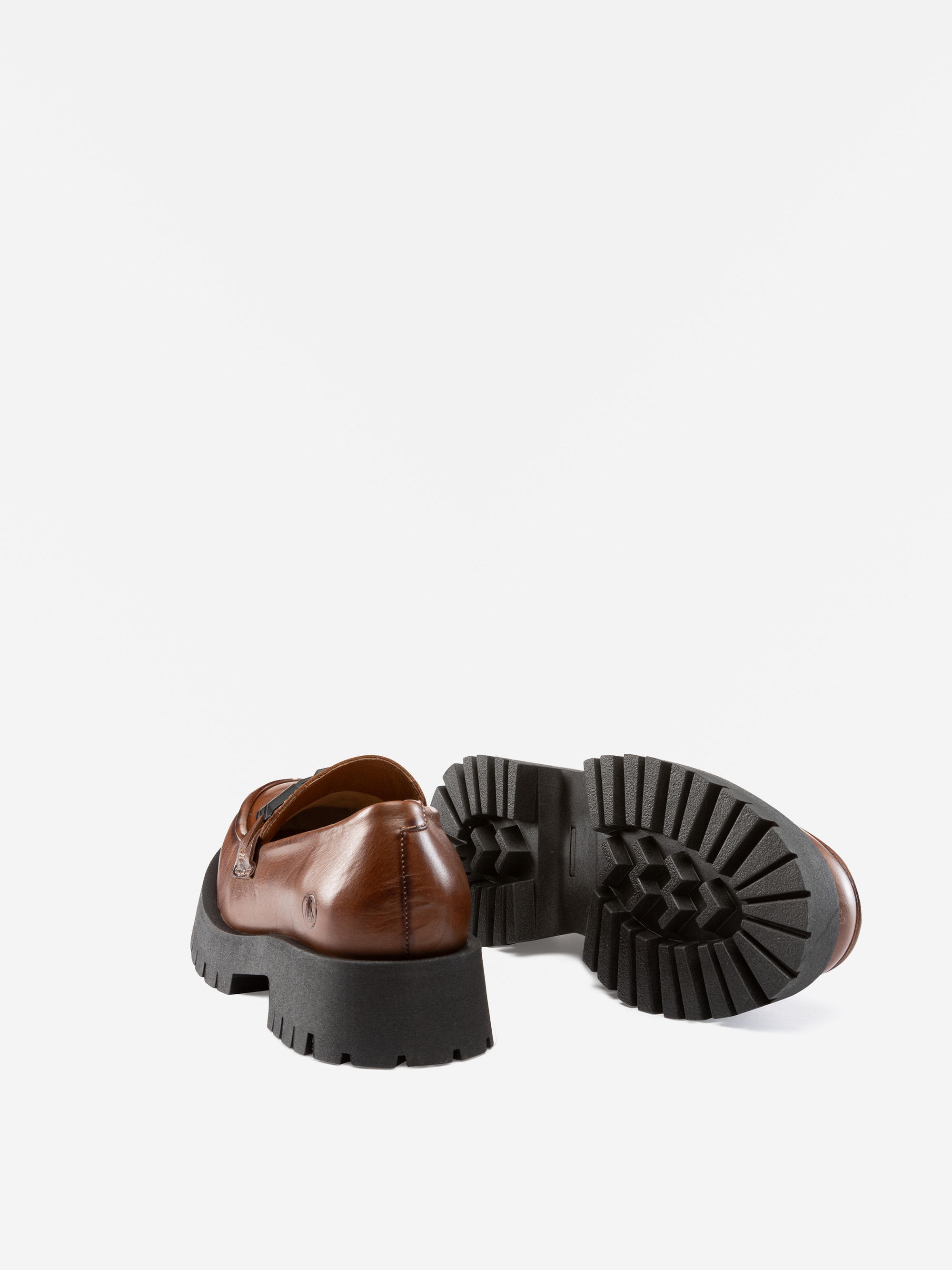 Ein Paar braune Lederslipper mit klobigen schwarzen Stollensohlen, wobei ein Schuh aufrecht steht und der andere auf der Seite liegt, um das Profil zu zeigen.