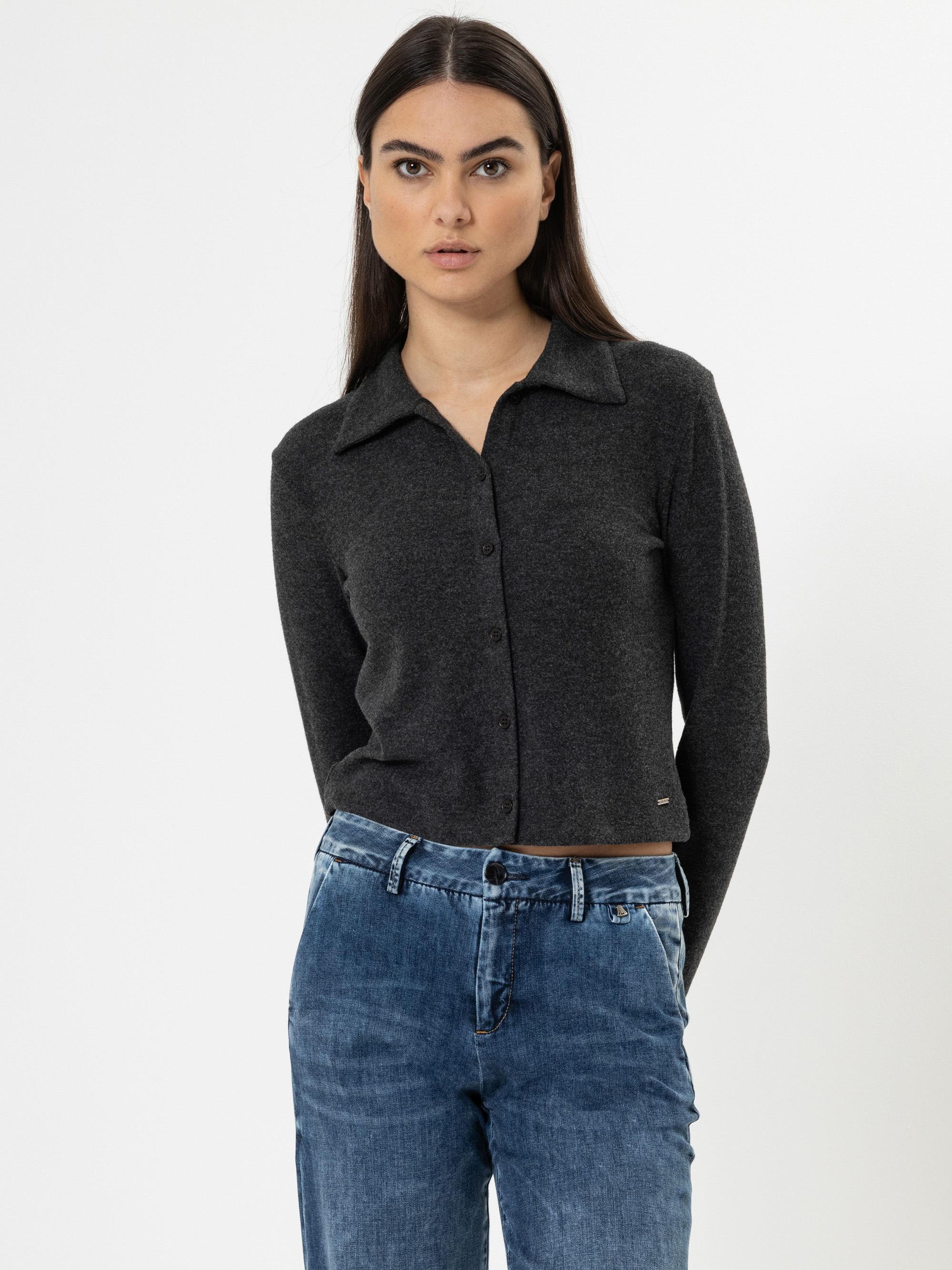 Eine Frau mit langen dunklen Haaren trägt eine dunkelgraue, hochgeknöpfte Strickjacke und blaue Jeans und steht vor einem schlichten weißen Hintergrund. Sie blickt direkt in die Kamera und hat einen neutralen Blick.