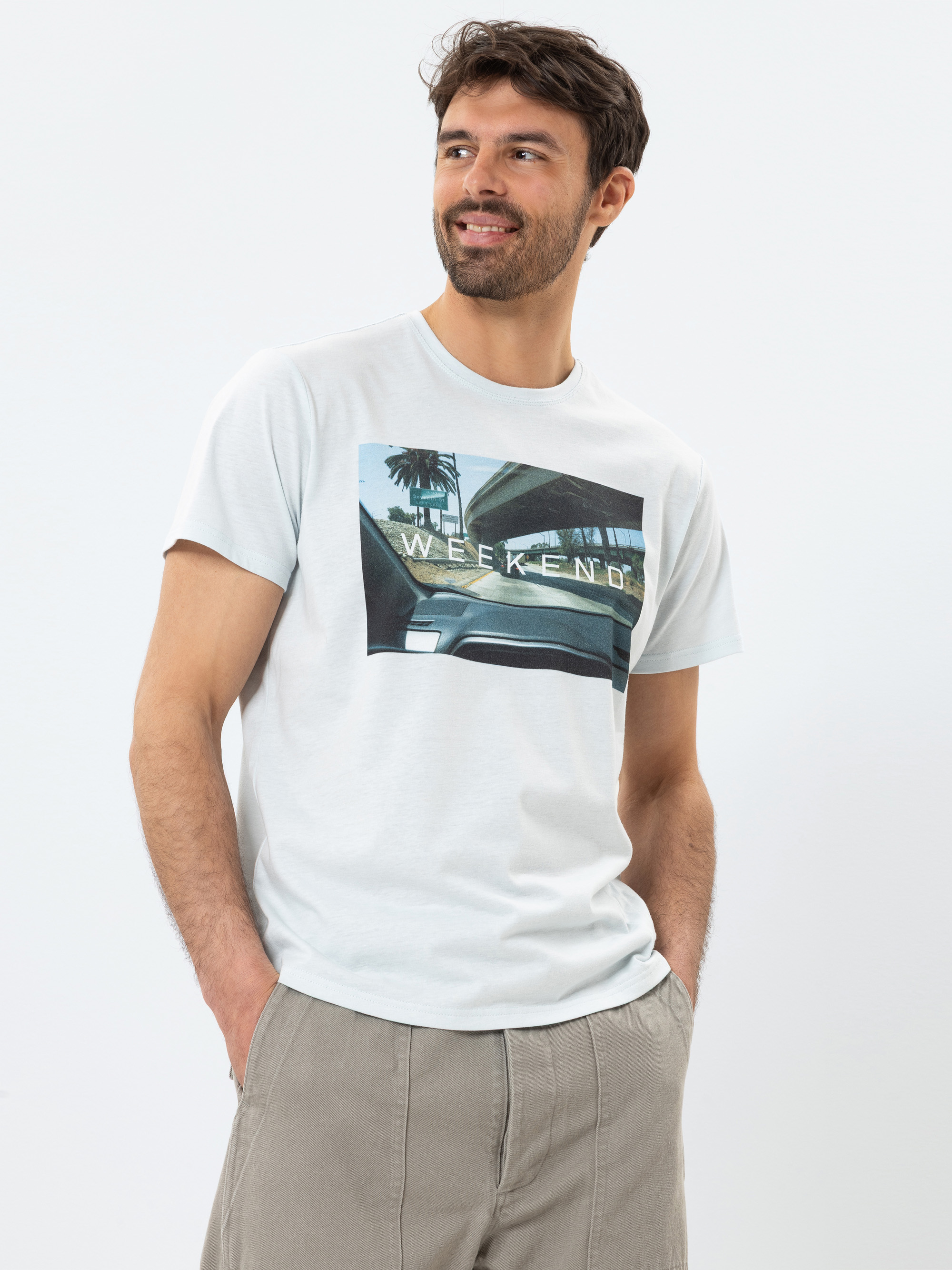 Ein Mann mit dunklem Haar und Bart, der ein hellgraues T-Shirt mit einer WEEKEND-Grafik und eine beigefarbene Hose trägt, steht mit einer Hand in der Hosentasche, lächelt und blickt zur Seite vor einem weißen Hintergrund.
