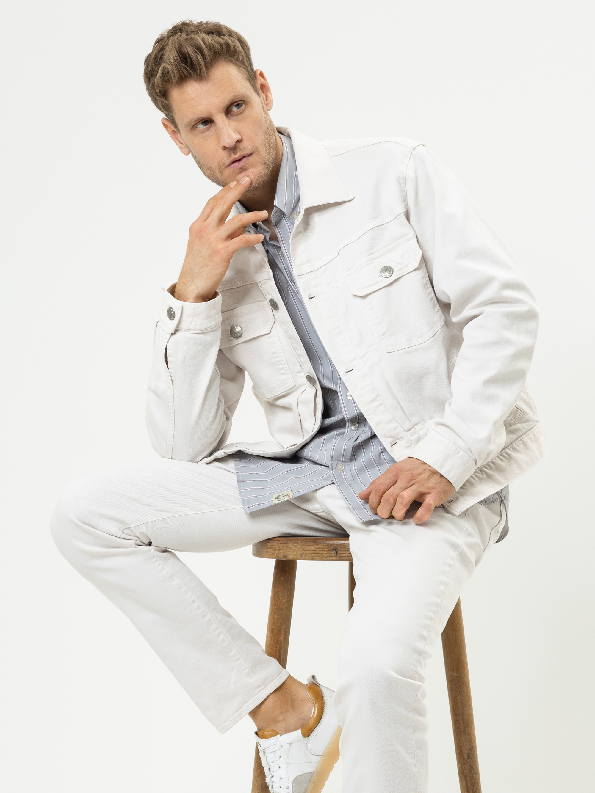 Ein Mann in weißer Jeansjacke, weißer Hose und weißen Turnschuhen sitzt auf einem Holzschemel, stützt sein Kinn auf die Hand und schaut nachdenklich. Er hat hellbraunes Haar und einen gestutzten Bart. Der Hintergrund ist einfarbig weiß.