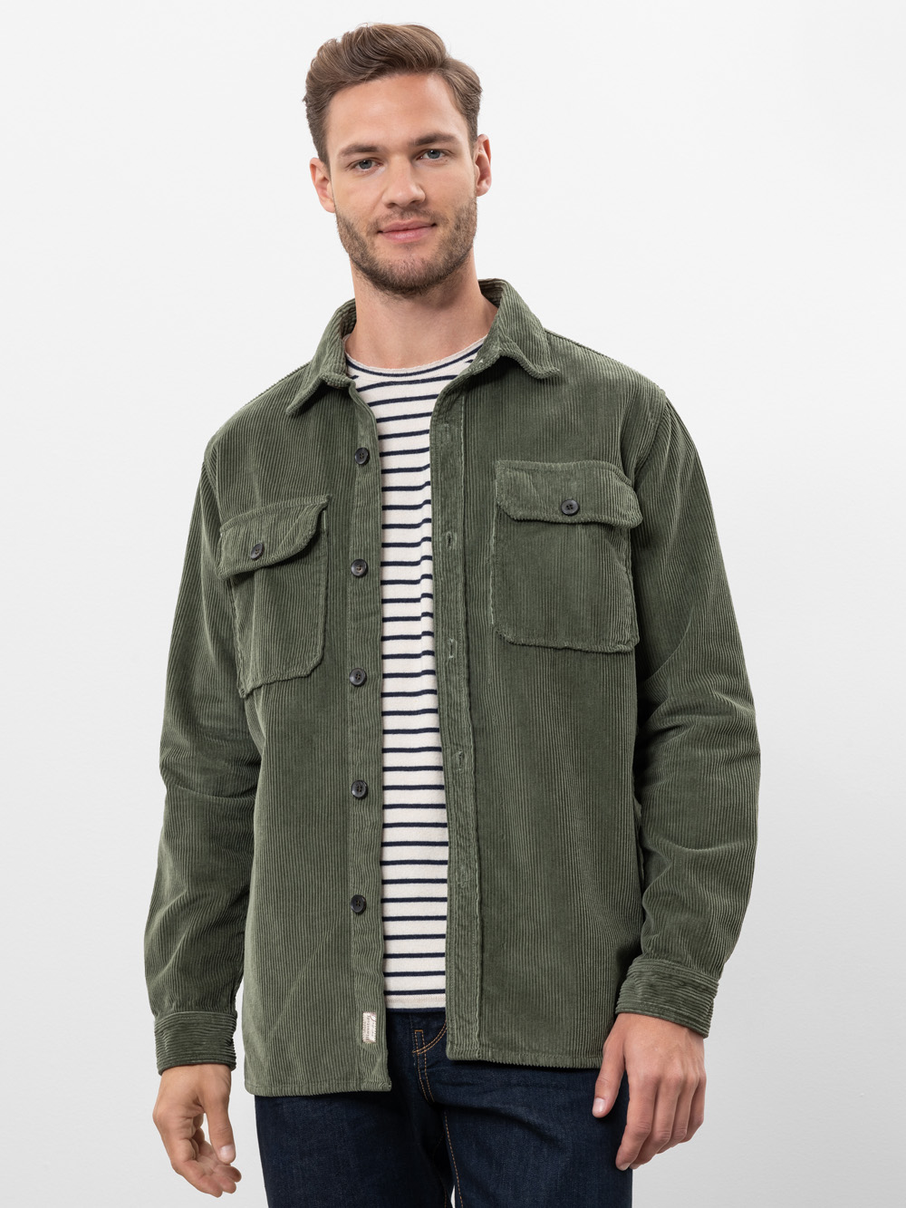 Ein Mann mit kurzen braunen Haaren, gestutztem Bart und leichtem Lächeln trägt ein olivgrünes Cord-Button-up-Hemd über einem schwarz-weiß gestreiften T-Shirt und eine dunkle Jeans, die vor einem schlichten weißen Hintergrund steht.