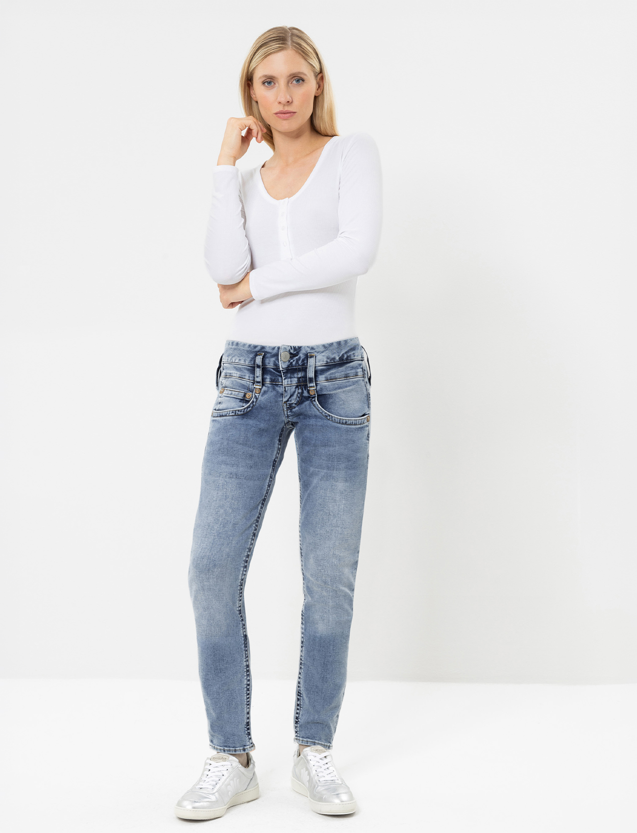 Eine Frau steht vor einem schlichten weißen Hintergrund und trägt eine ärmellose Jeansweste, Blue Skinny Jeans und dunkelblaue Slide-Sandalen. Sie hat langes glattes Haar und einen neutralen Gesichtsausdruck.