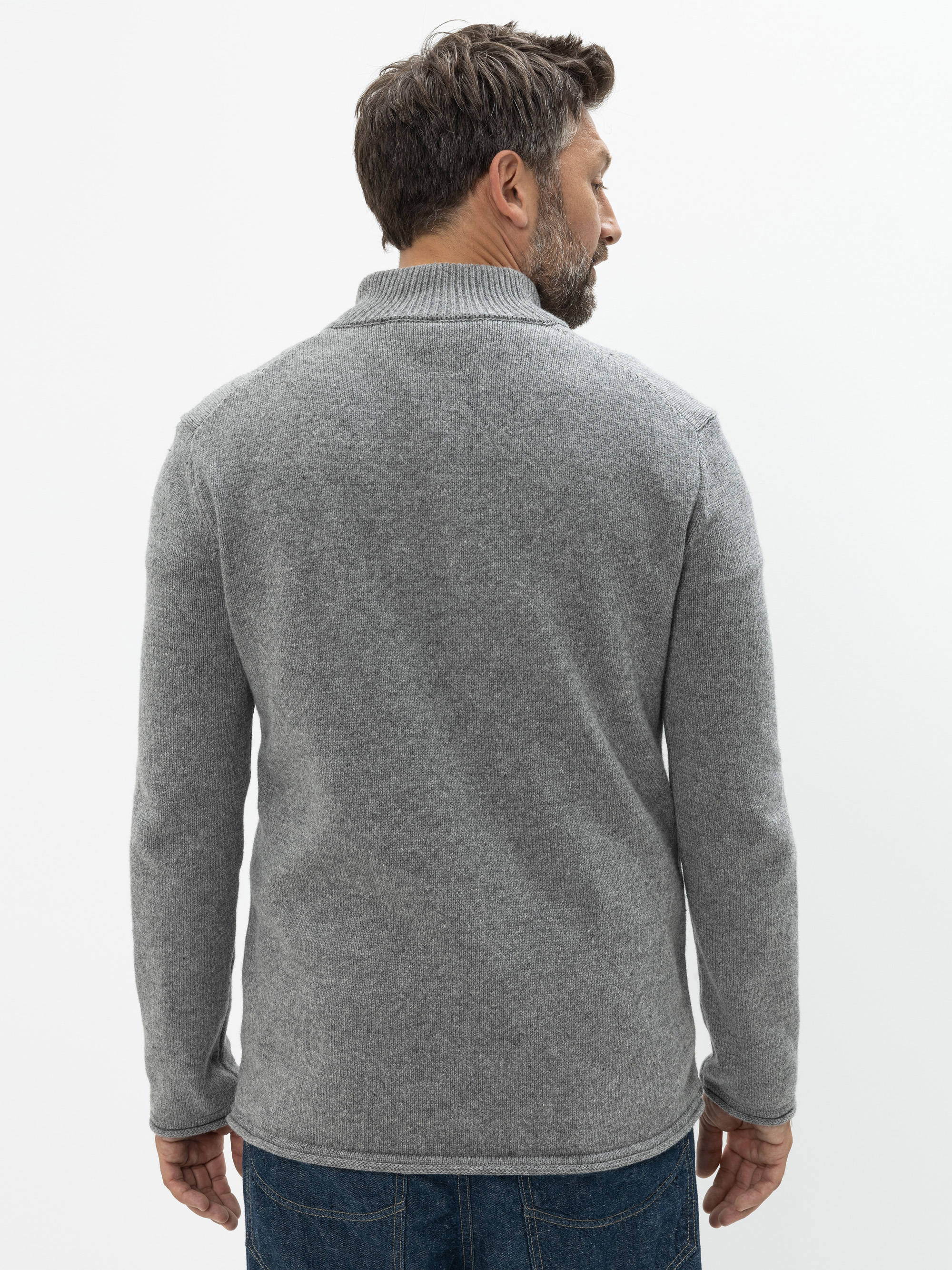 Ein Mann mit kurzen dunklen Haaren und einem Bart steht mit dem Rücken zur Kamera, trägt einen grauen Strickpullover und blaue Jeans vor einem schlichten weißen Hintergrund.