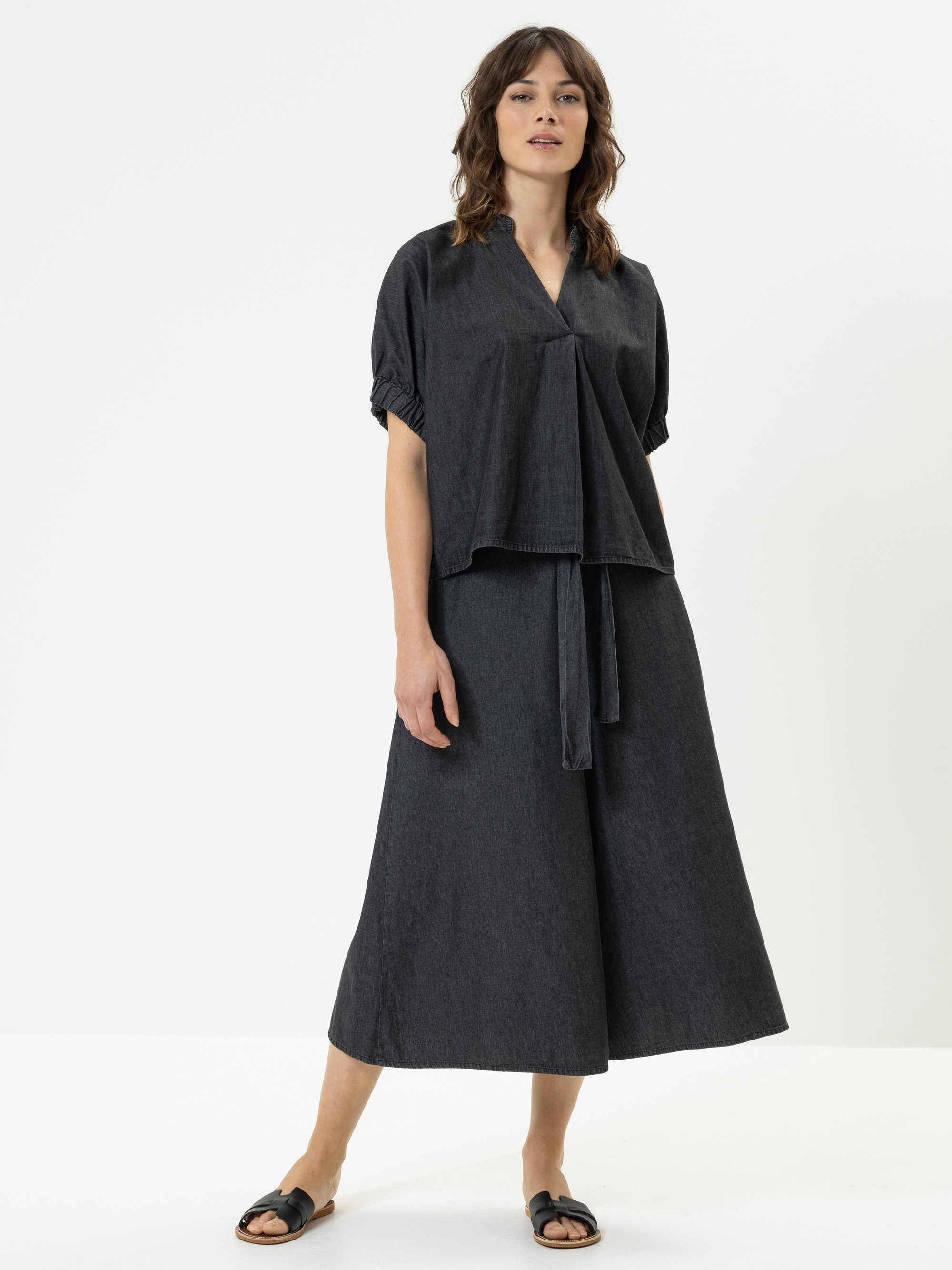 Eine Frau steht vor einem weißen Hintergrund und trägt ein locker sitzendes, dunkelgraues Oberteil mit kurzen Ärmeln und einen passenden, weit geschnittenen Rock. Sie trägt schwarze Slide-Sandalen und hat gewelltes braunes Haar.