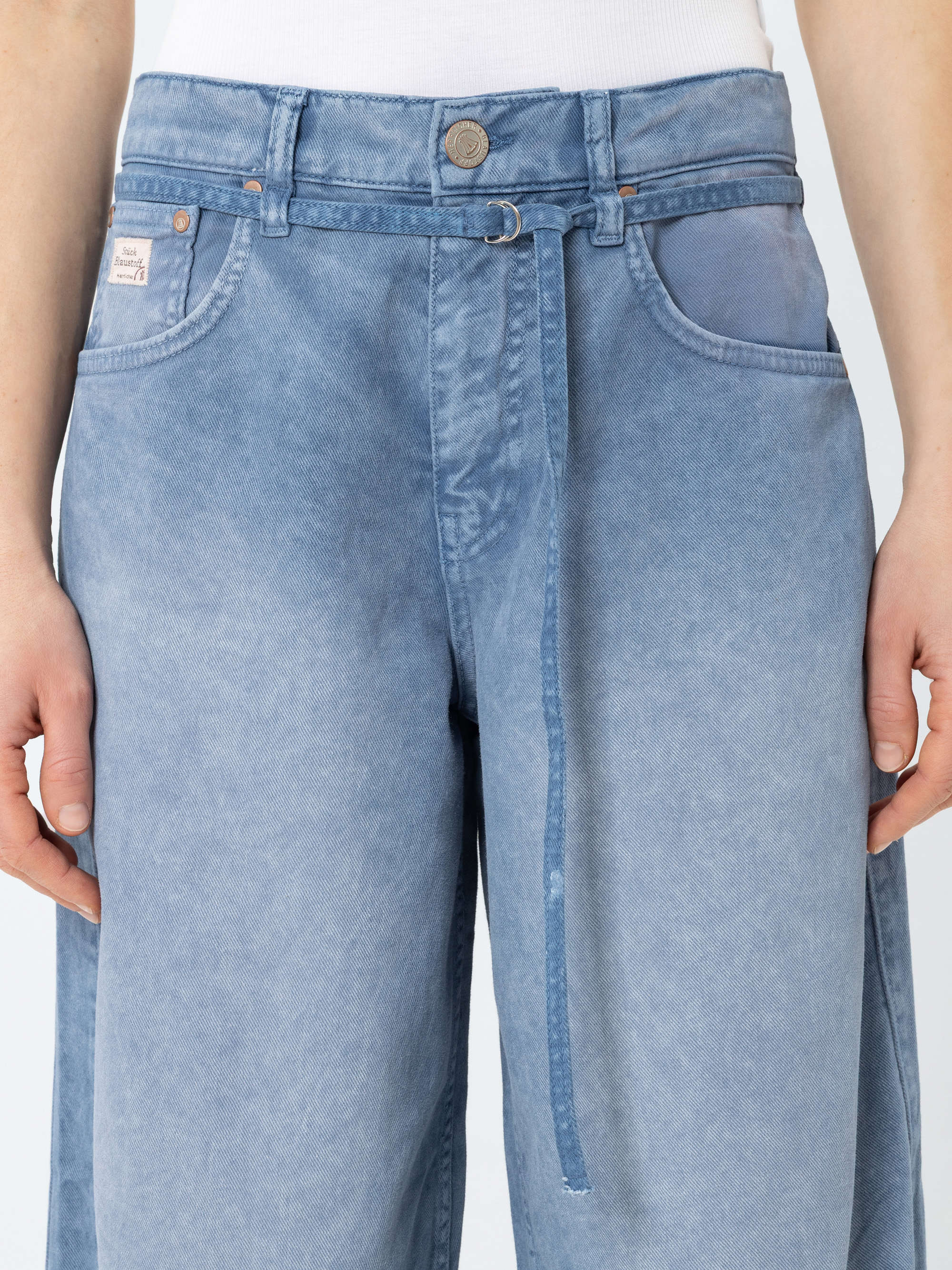 Eine Person, die eine hellblaue Jeans mit weitem Bein und einer Krawatte in der Taille und einem Knopfverschluss trägt, steht vor einem unifarbenen Hintergrund; nur der Oberkörper und die Oberschenkel sind sichtbar.